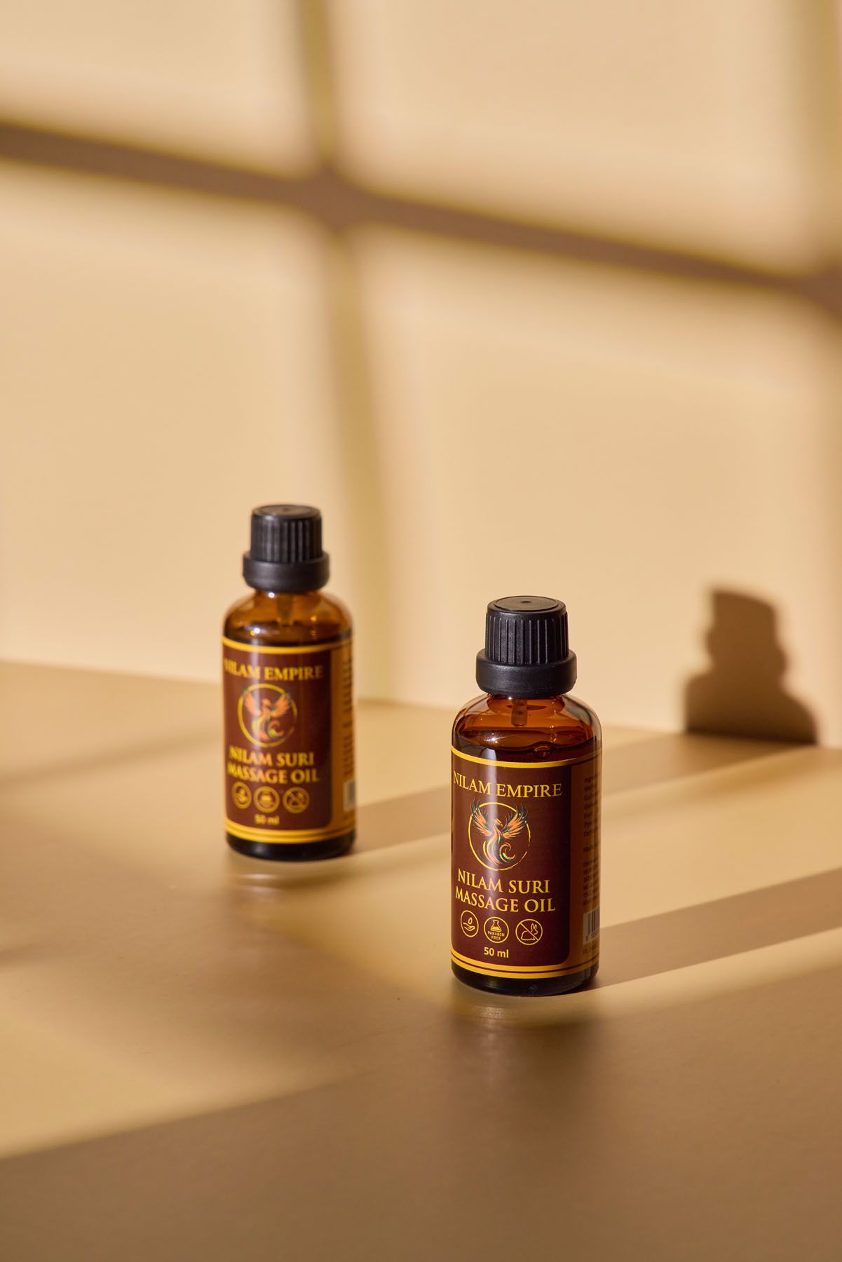 Two Hilam Empire Urut Suri massage oil amber bottles displayed on a warm beige surface with window shadows.