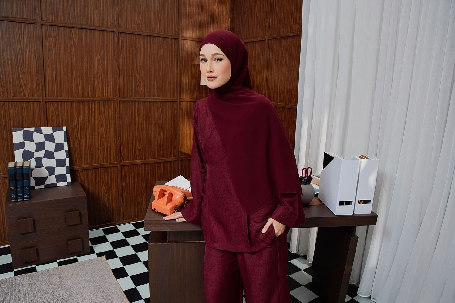 Woman in burgundy hijab and matching outfit holds an orange object in a stylish checkered floor room.
