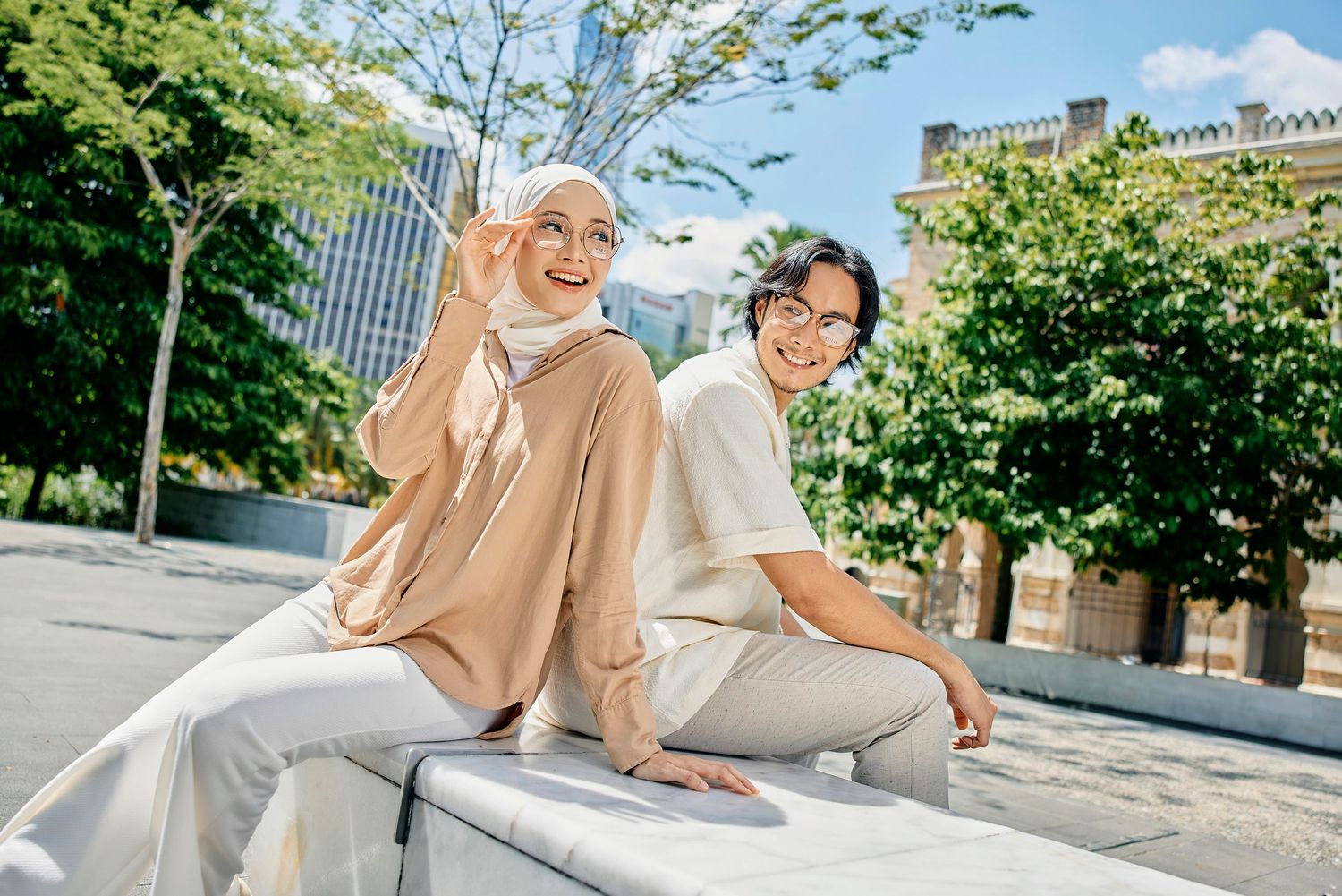 Two women sitting on an outdoor bench, one wearing a hijab, smiling in a sunny urban setting with green trees.