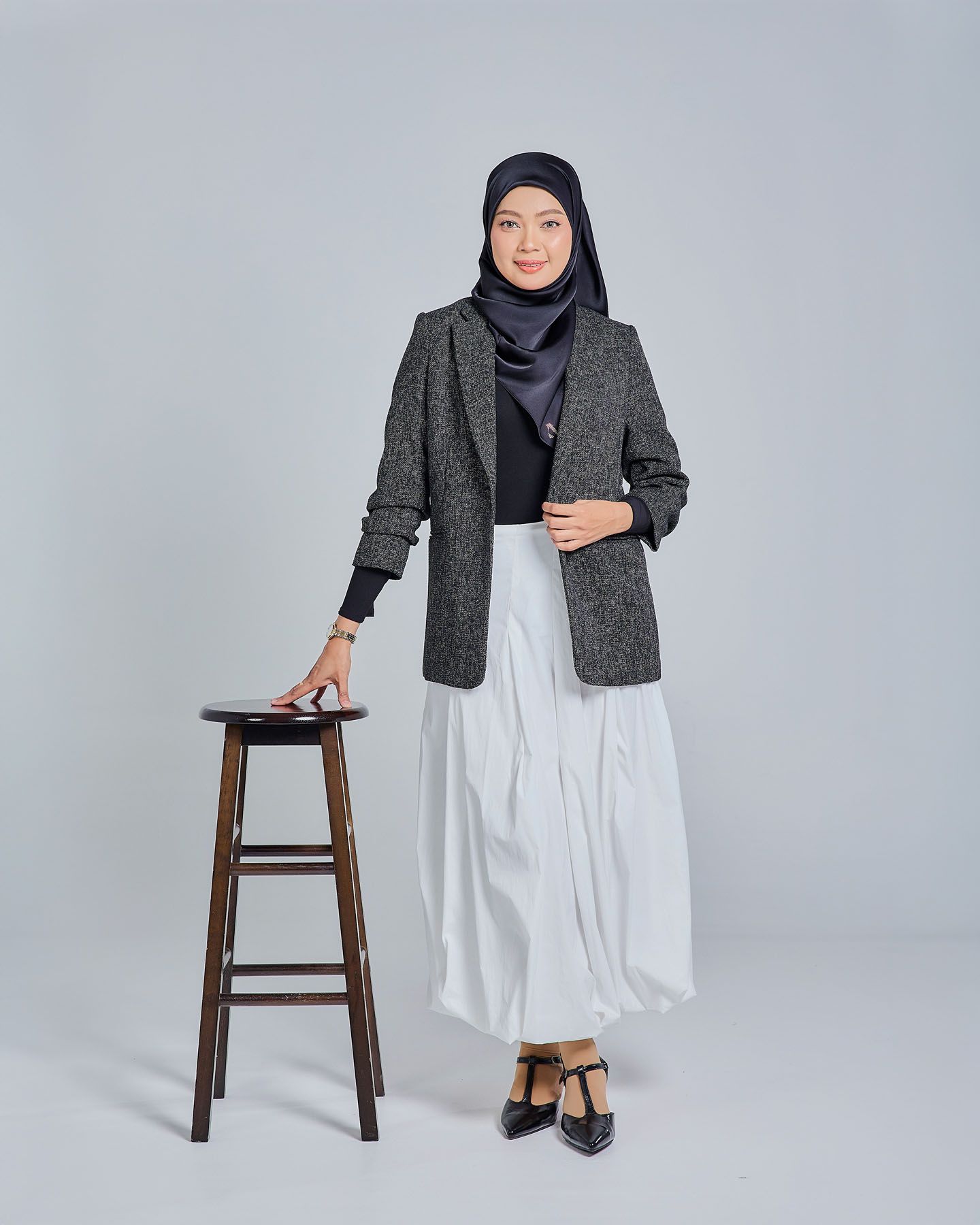 Muslim woman in gray herringbone blazer, white skirt, and navy hijab posing beside a wooden stool.
