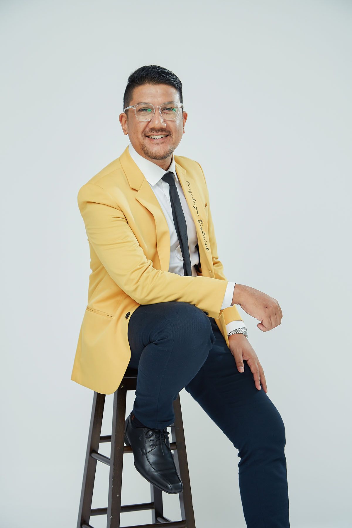 Smiling Asian man wearing glasses and yellow blazer sitting relaxed on wooden stool in studio.