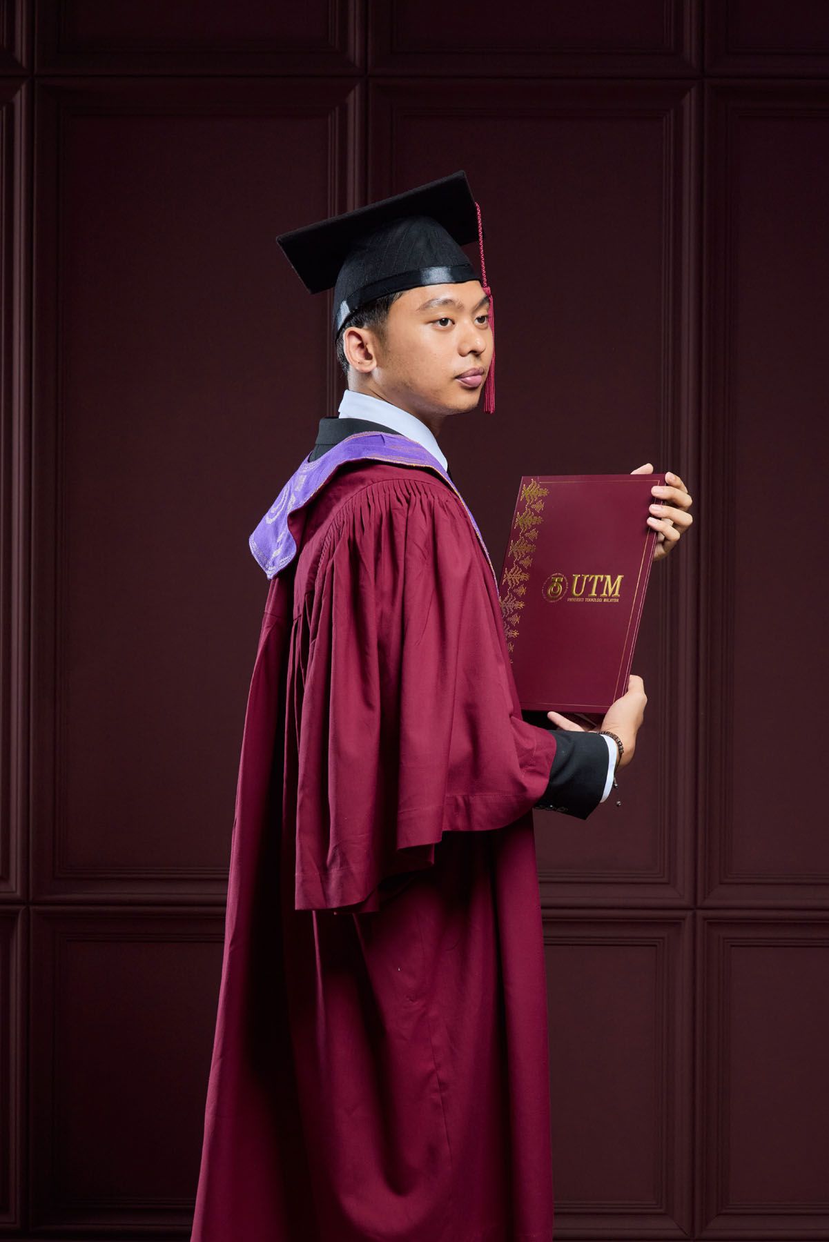 Side profile of male graduate in maroon UTM graduation gown holding university diploma certificate.