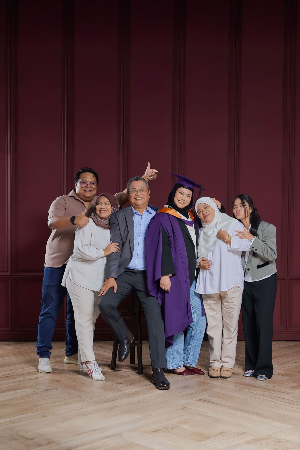 Family celebrating graduation with woman in purple cap and gown against dark red wall.