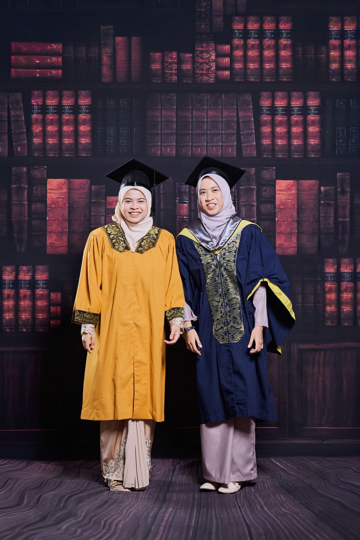 Two Muslim women graduates in golden and navy blue robes smiling at graduation ceremony.