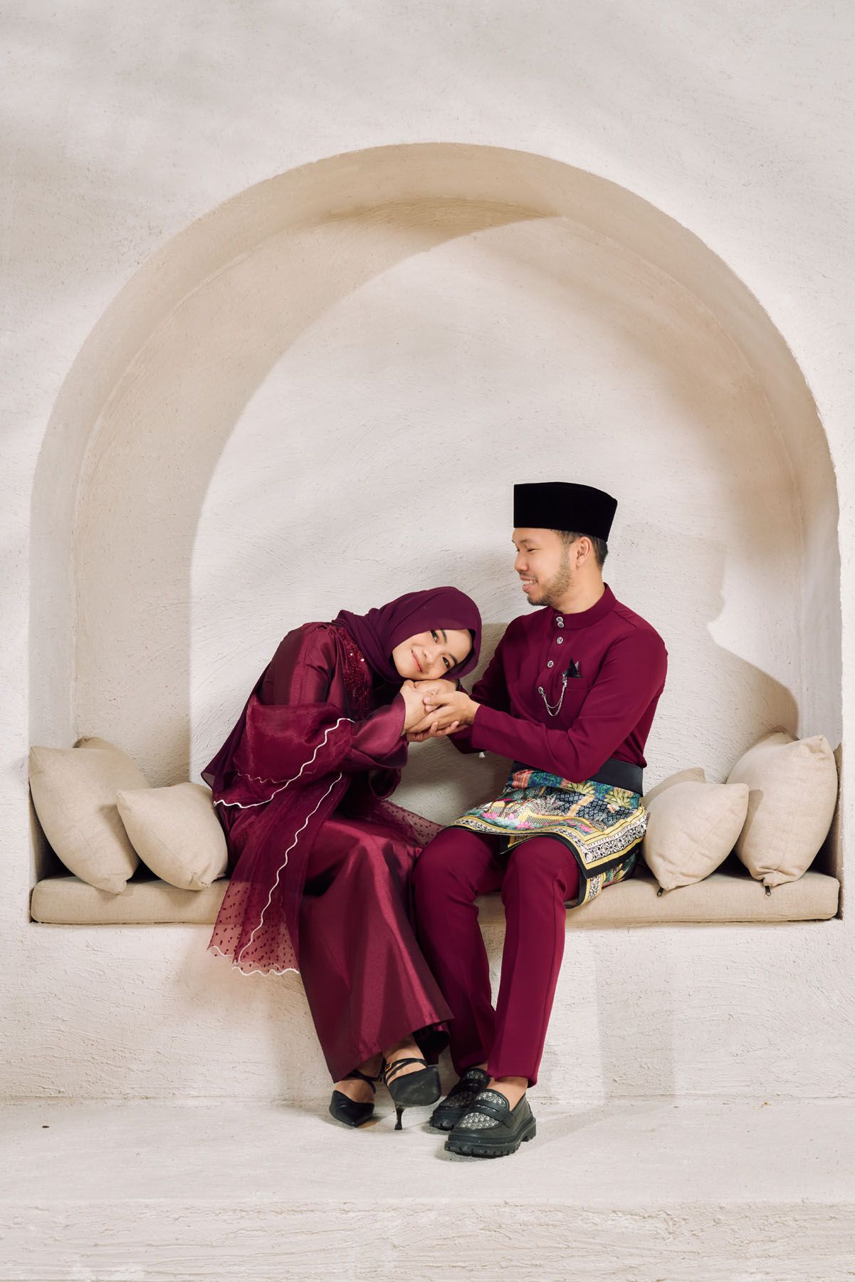 A romantic Malaysian couple in matching burgundy traditional outfits sitting together under a white arched alcove.
