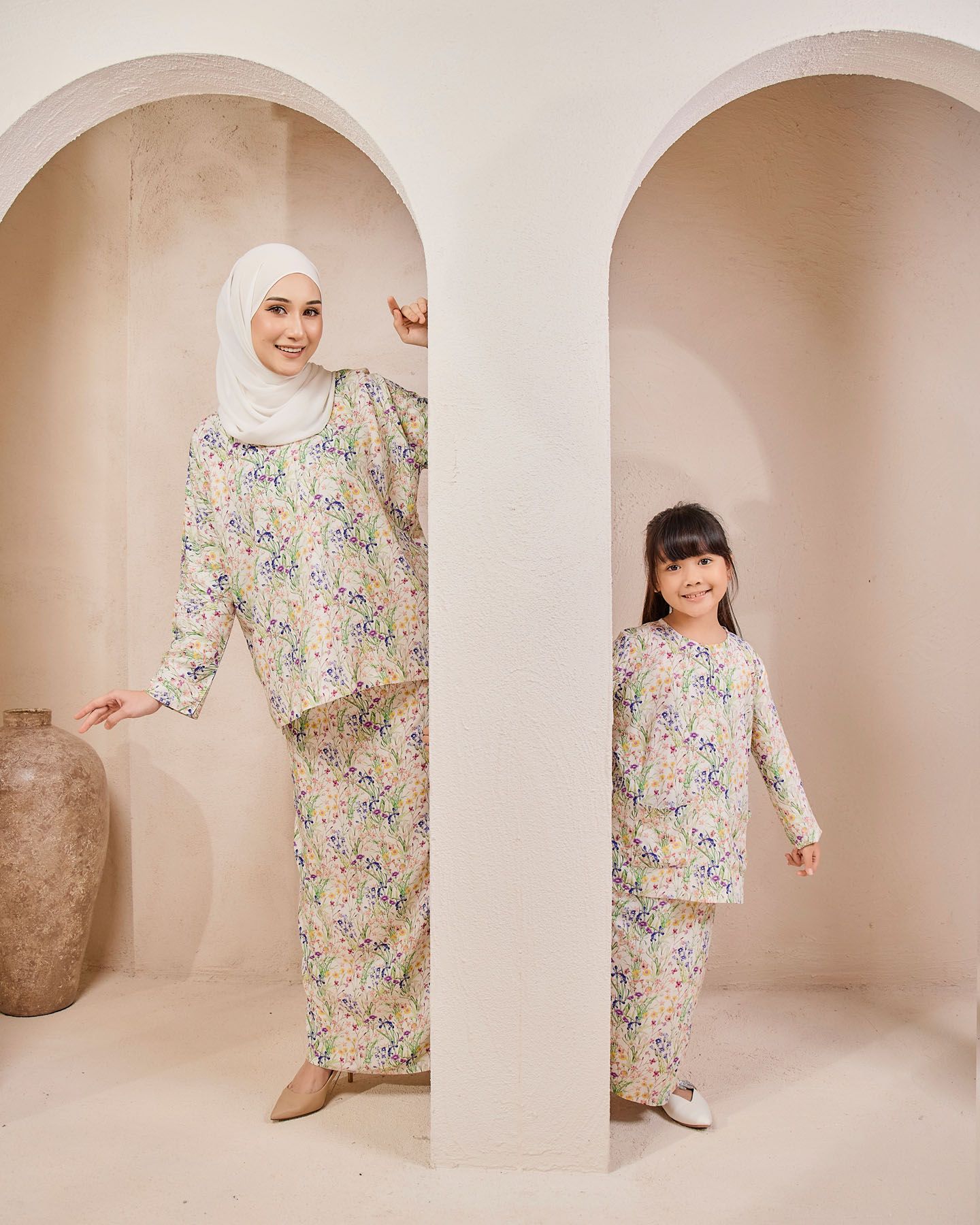 A mother and daughter in matching floral baju kurung outfits pose on opposite sides of a white archway pillar.