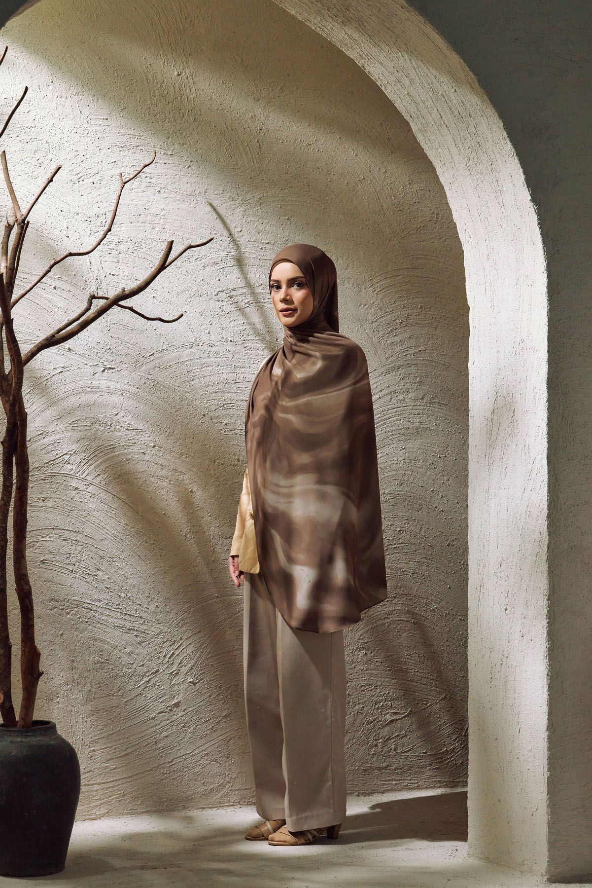 Muslim woman wearing a brown abstract-print hijab and matching outfit standing in a white arched doorway.