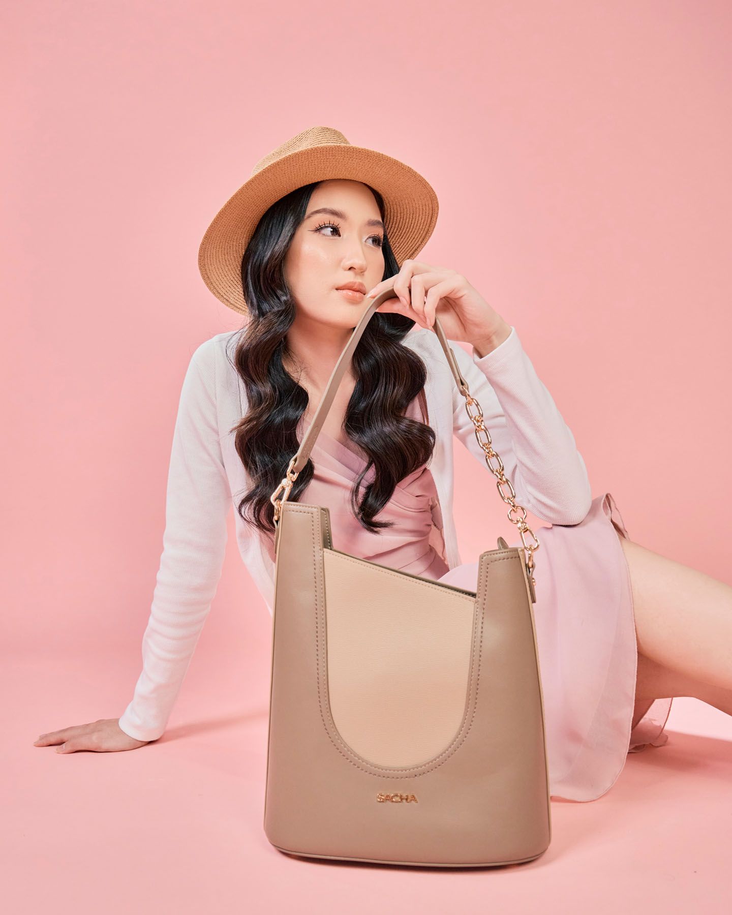 Asian woman in pink outfit sitting with beige bucket bag and gold chain strap.