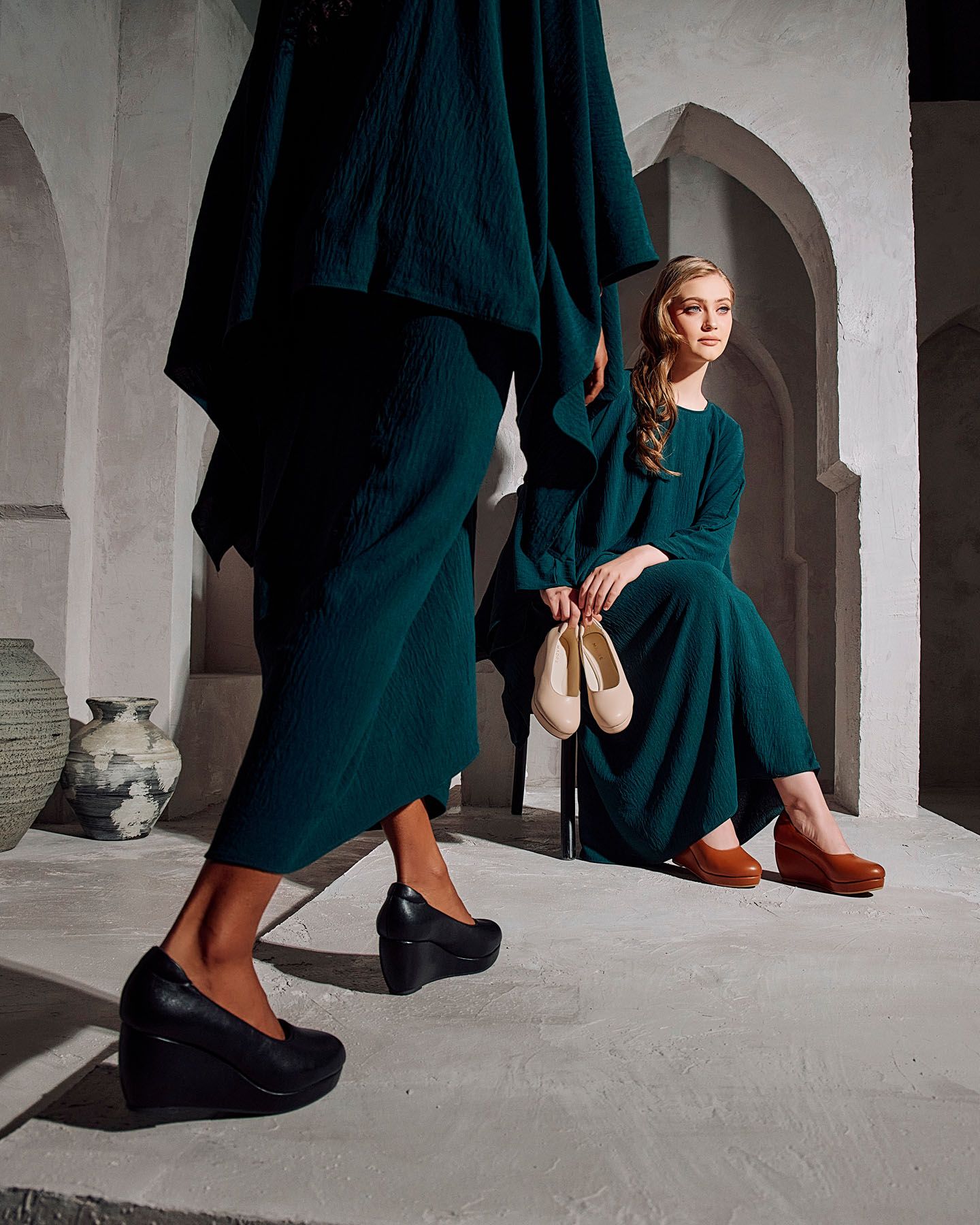 Two women in teal green dresses showcase black and brown wedge shoes in stone arch setting.