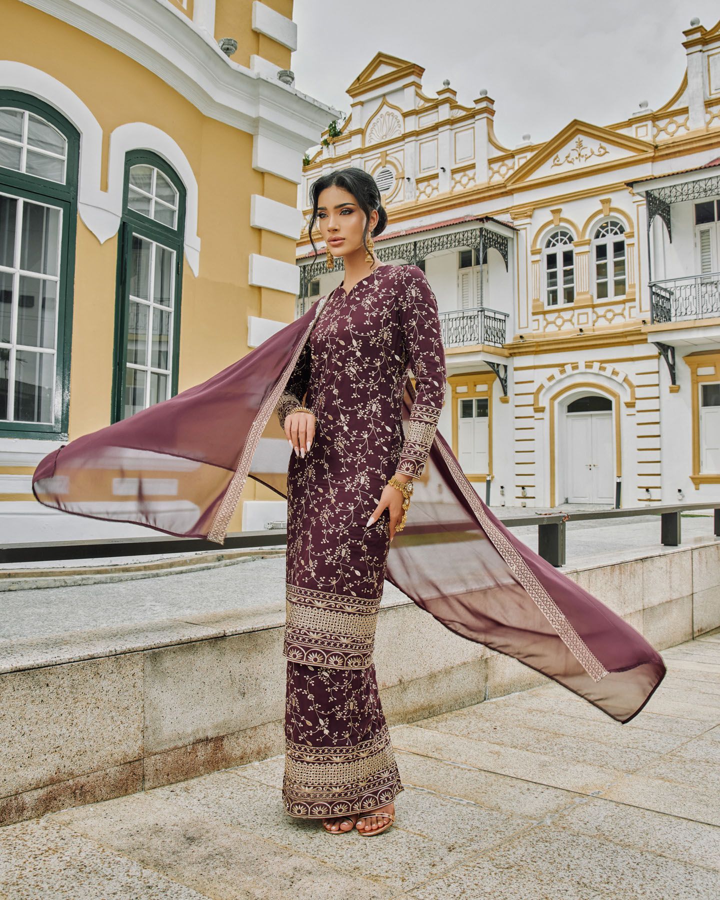 Woman in purple floral baju kurung with flowing cape poses before a colorful heritage building.