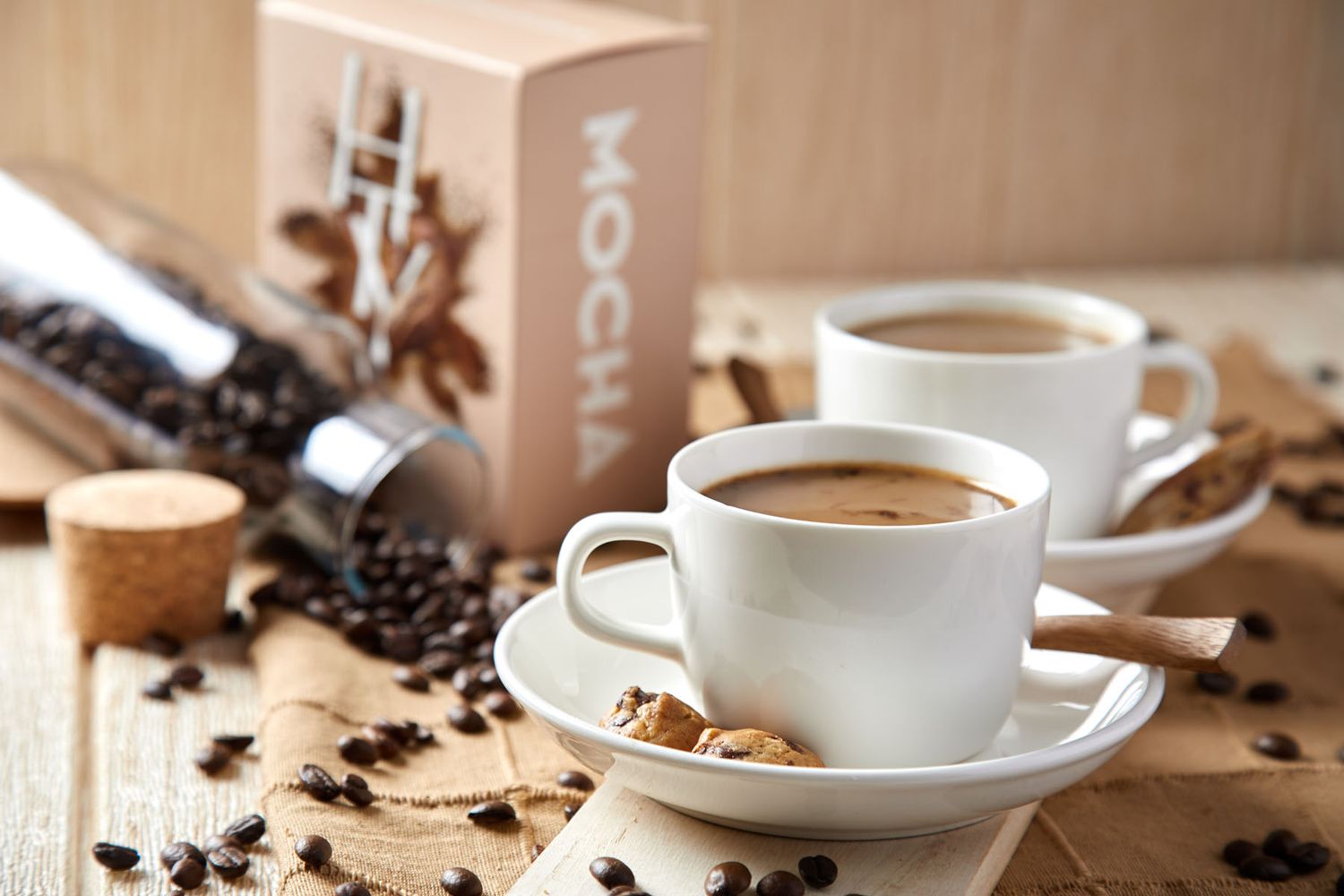 Two white cups of mocha coffee with coffee beans, brown sugar, and Mocha branded box on wooden surface.