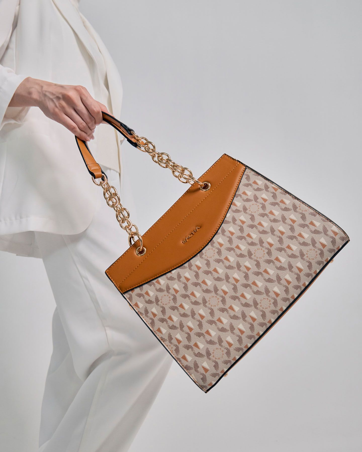 Woman in white outfit carrying a tan monogram tote bag with gold chain handles.