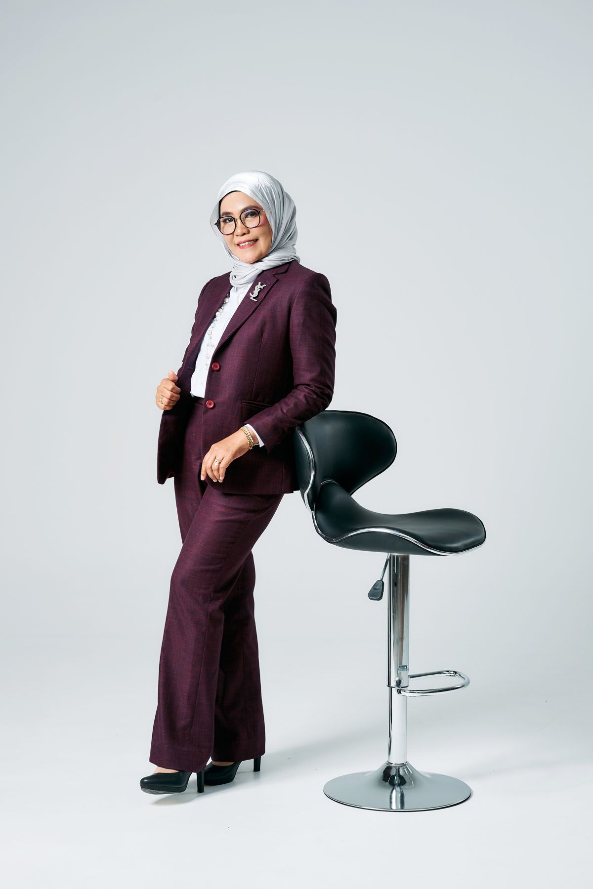 A professional Muslim woman in a burgundy suit and white hijab poses confidently beside a modern bar stool on a white background.
