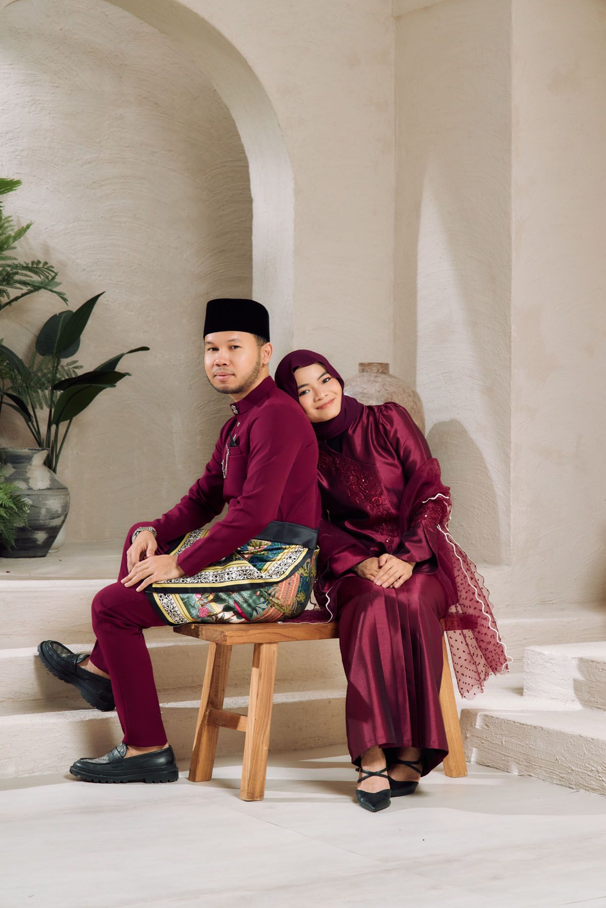 A Malaysian couple in coordinating deep red traditional Eid attire sitting back-to-back on a wooden bench indoors.
