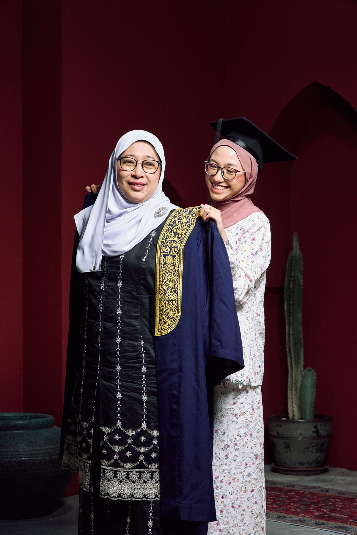 Two Muslim women smile together, one in graduation gown and mortarboard at ceremony.