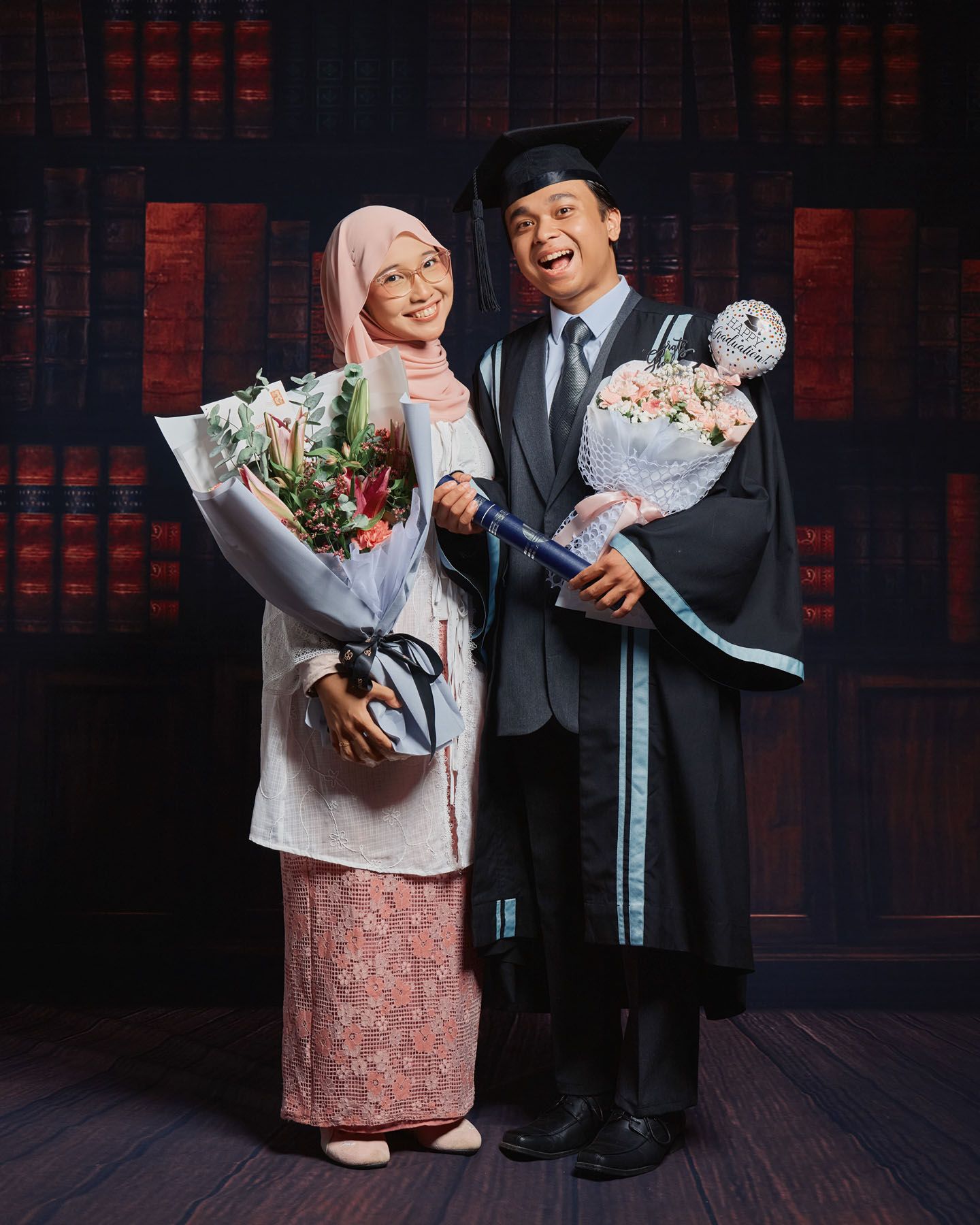Smiling graduate in cap and gown holding bouquet with companion in pink hijab and floral skirt.