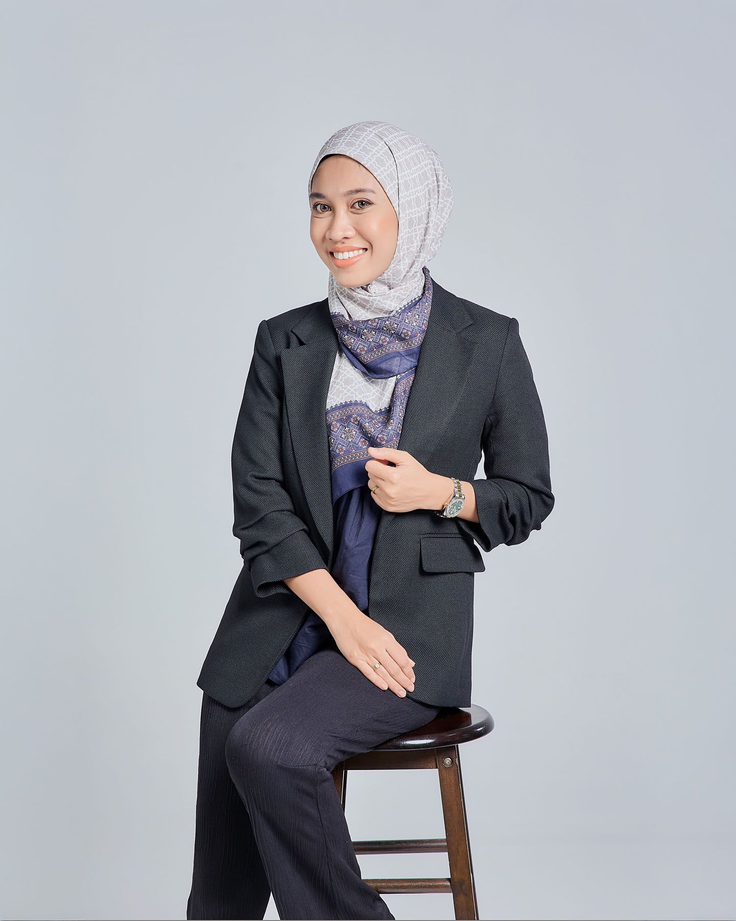 Smiling Muslim woman in dark blazer and navy scarf with gray hijab seated on wooden stool.