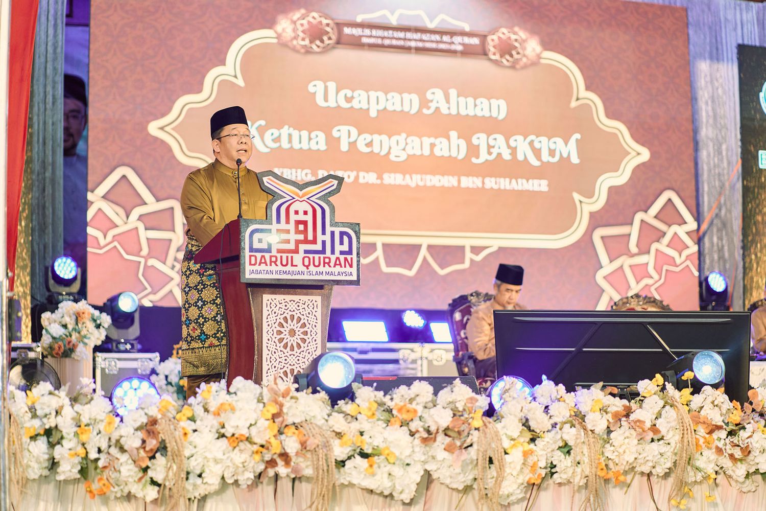 Malaysian official in traditional Baju Melayu delivers speech at a Darul Quran JAKIM formal ceremony.