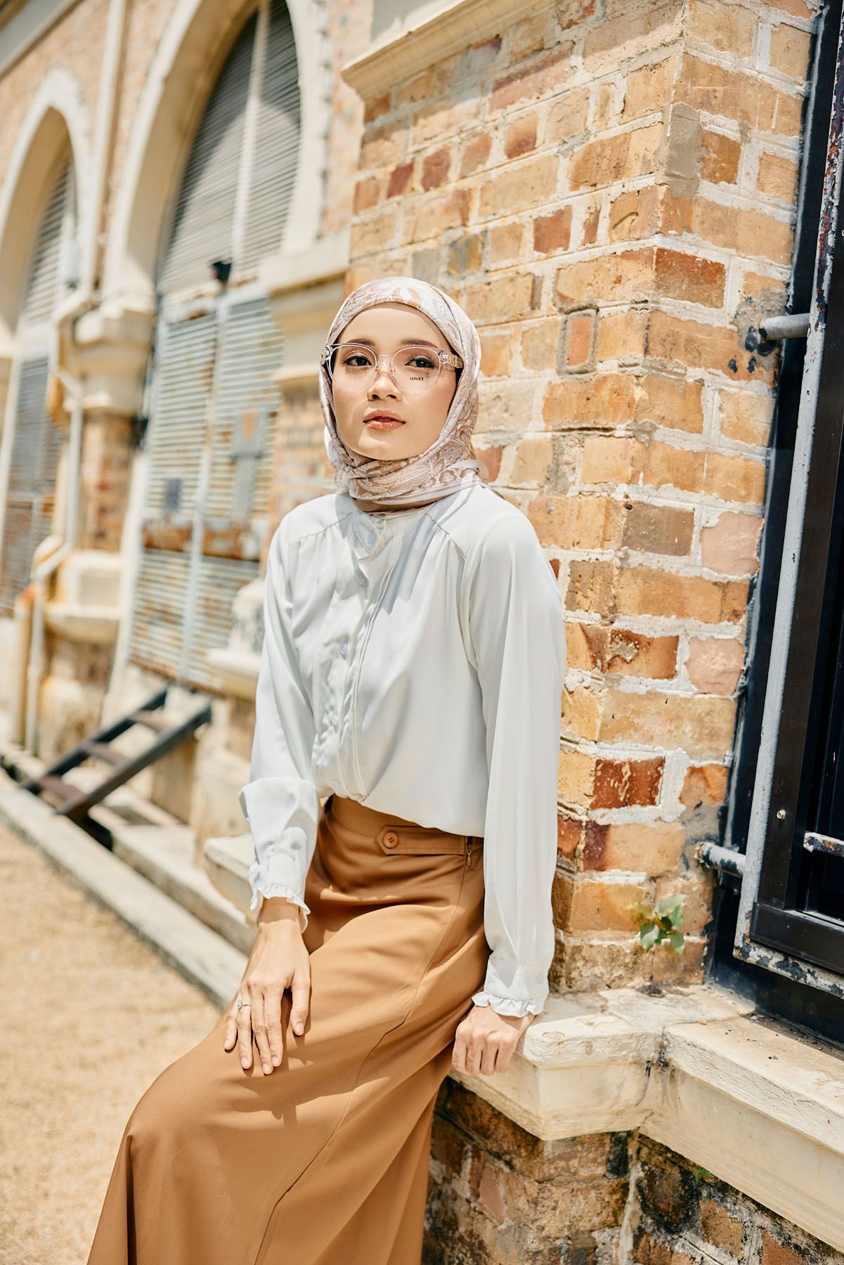 Woman in white blouse and brown skirt wearing glasses and hijab by brick wall.