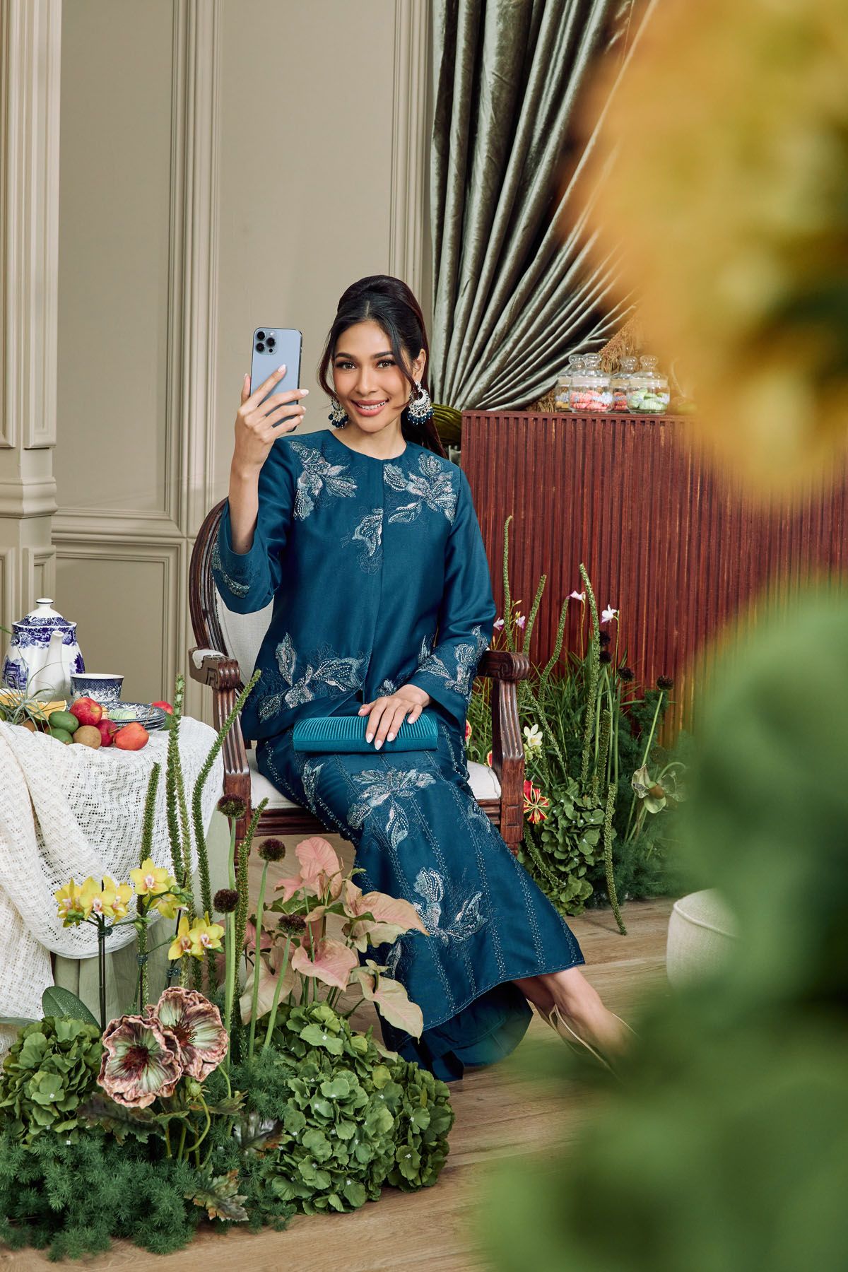 Woman in teal floral baju kurung taking a selfie while seated among lush green plants in a decorated room.