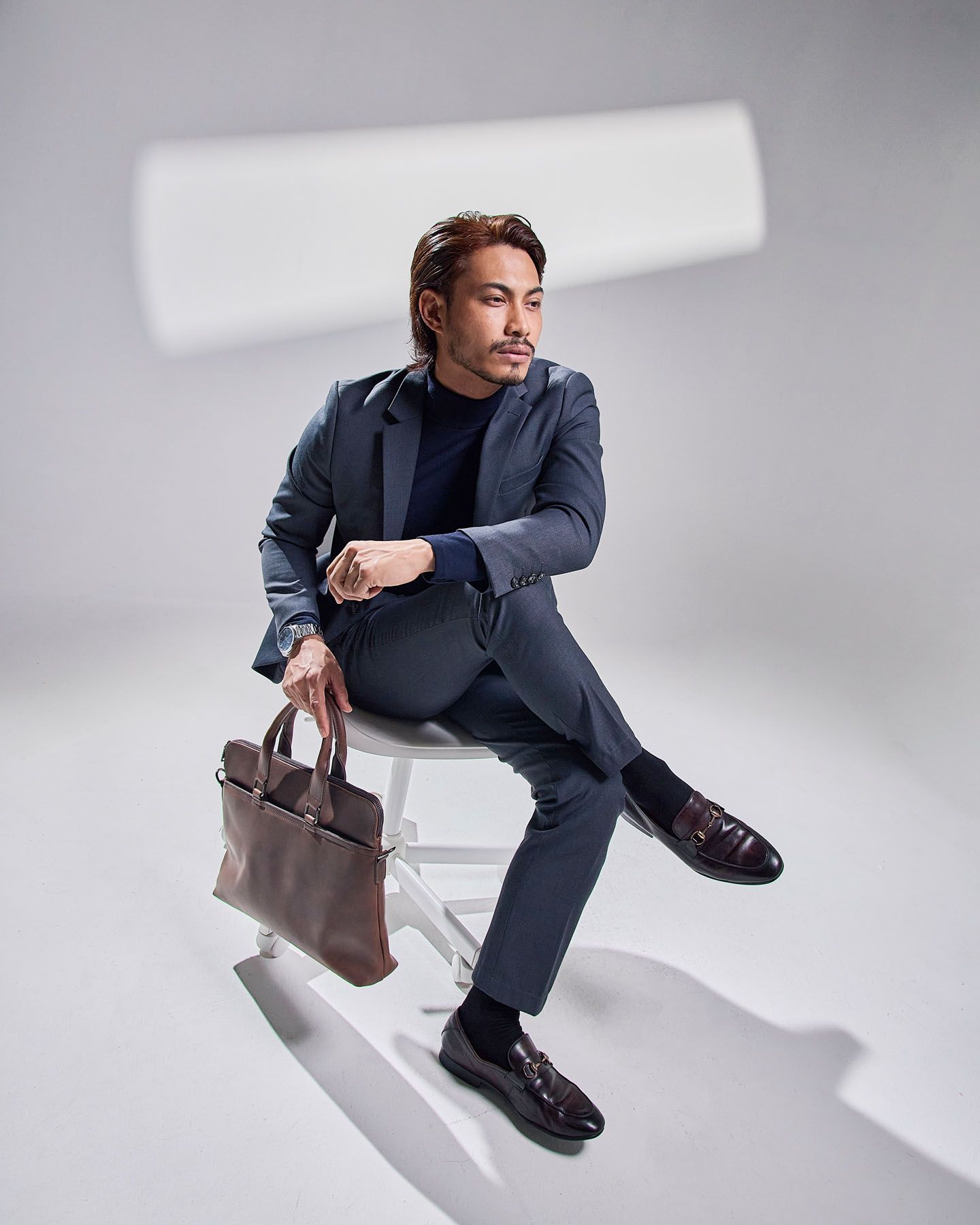 Fashionable man in grey suit holding brown leather bag seated on white stool against light background.
