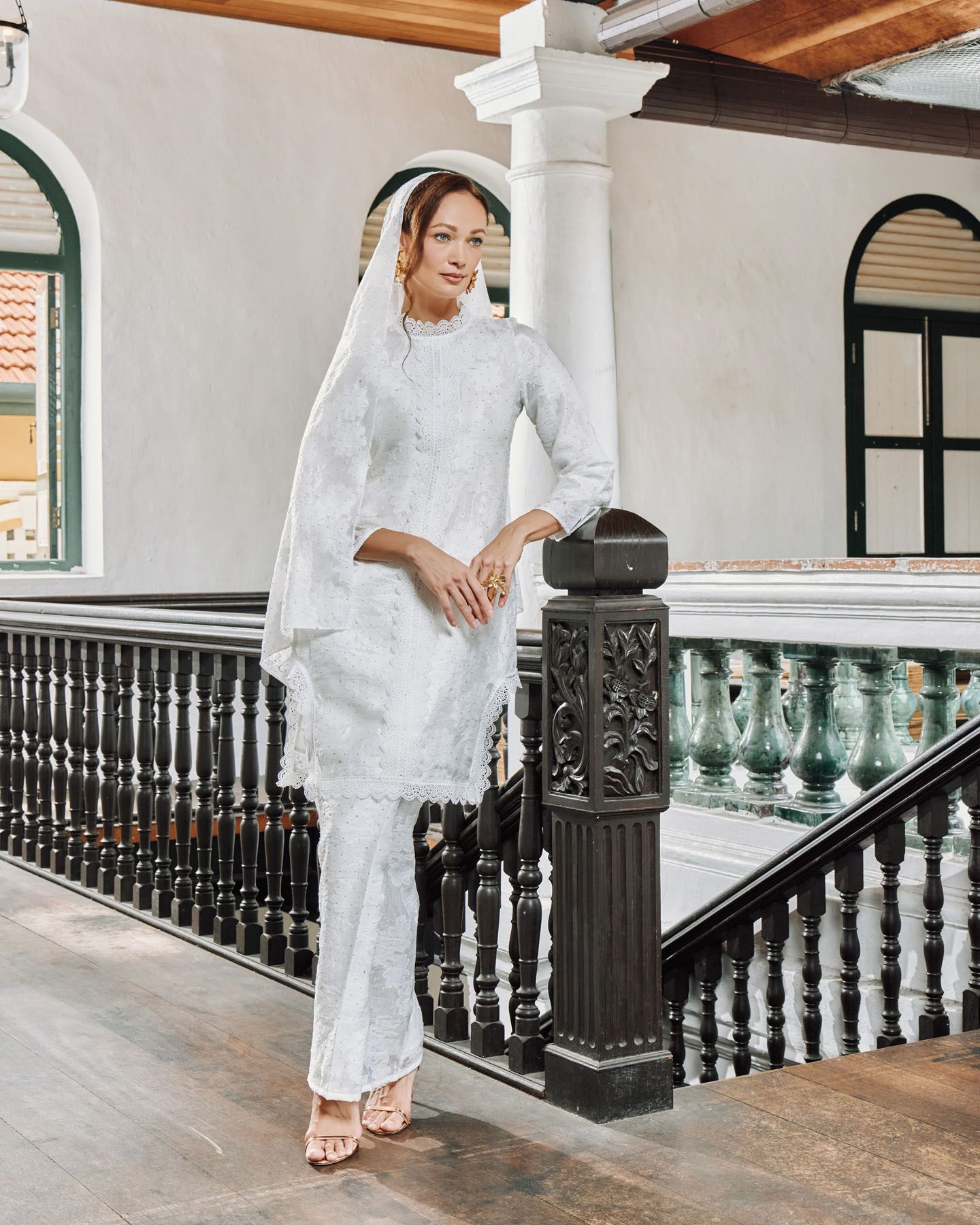 Woman in white lace baju kurung with veil stands on a heritage building staircase.