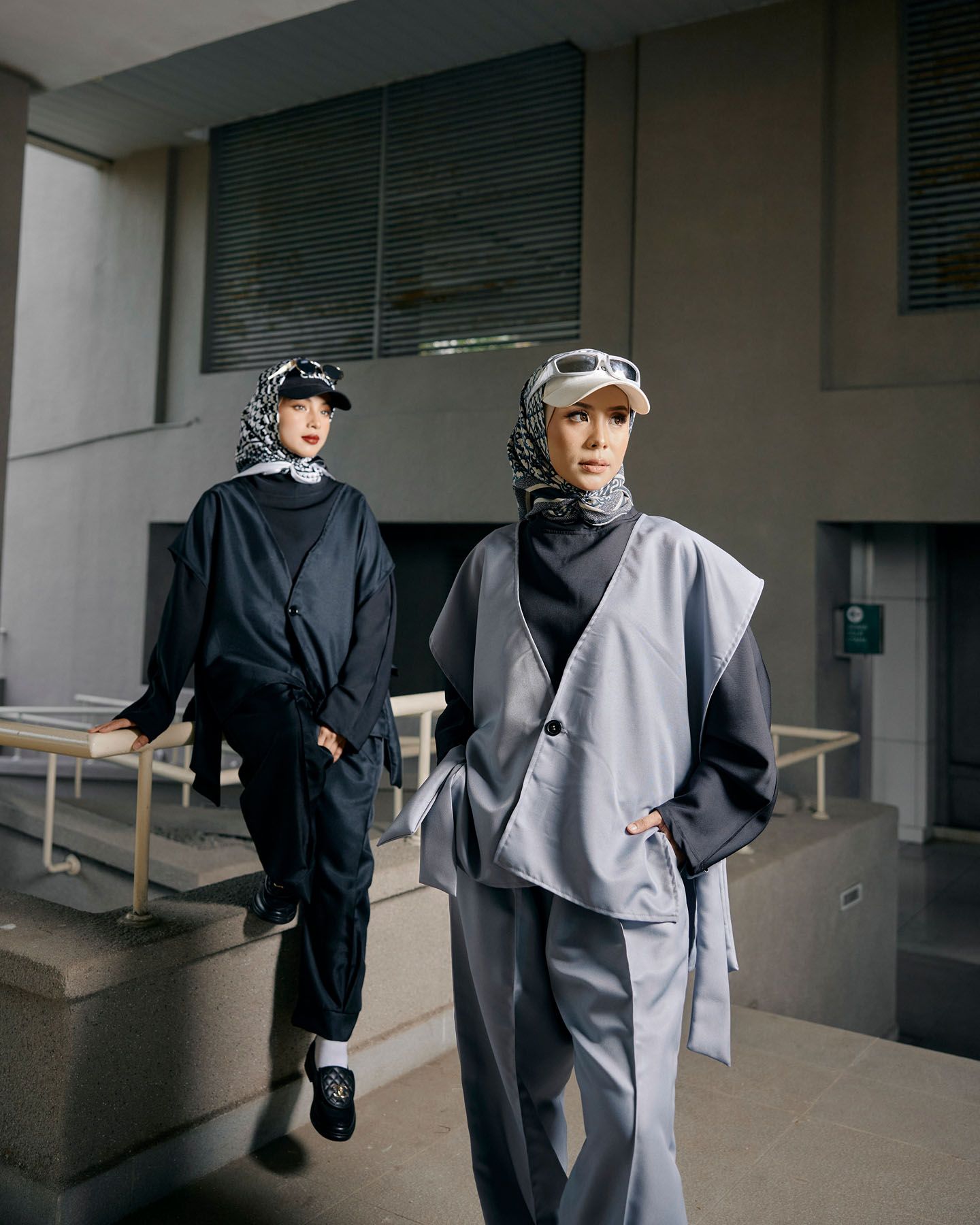 Two women in coordinated grey and black outfits with patterned hijabs walking in urban setting.