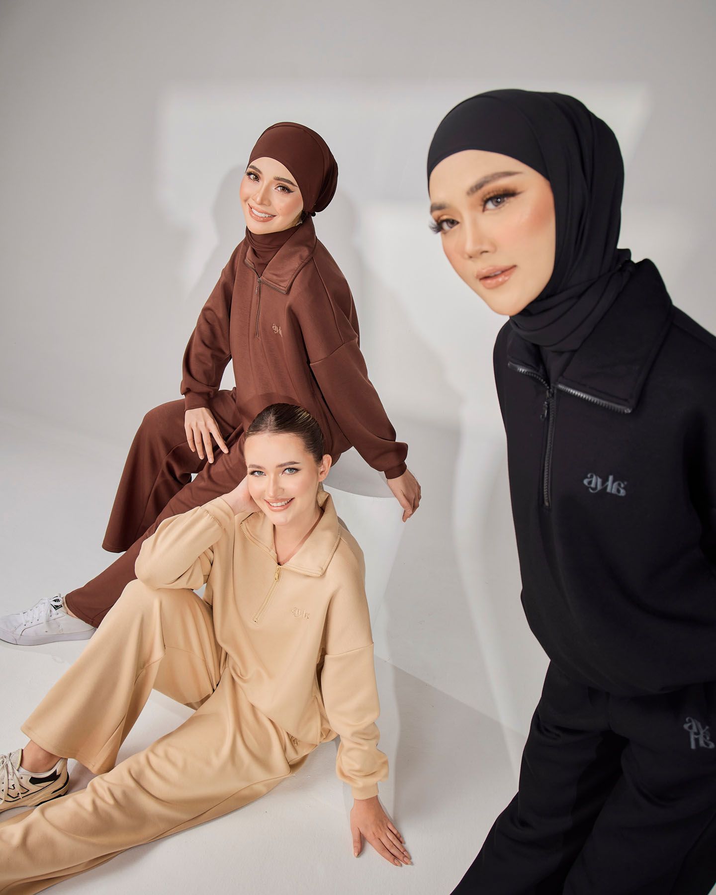 Three women model matching tracksuit sets in black, brown, and beige with hijab styling.
