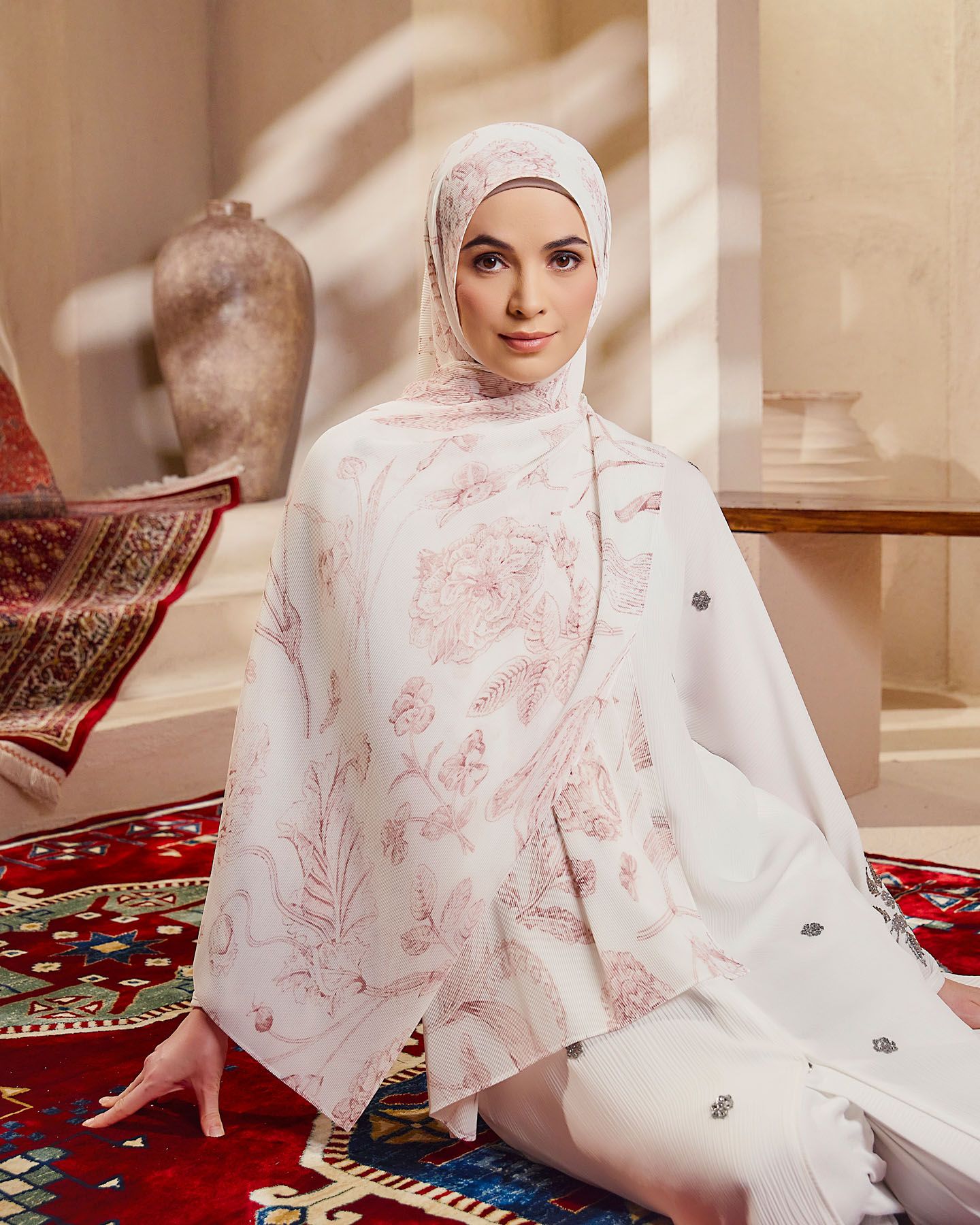 Elegant woman in white floral hijab and outfit seated on a colorful rug in a styled room.