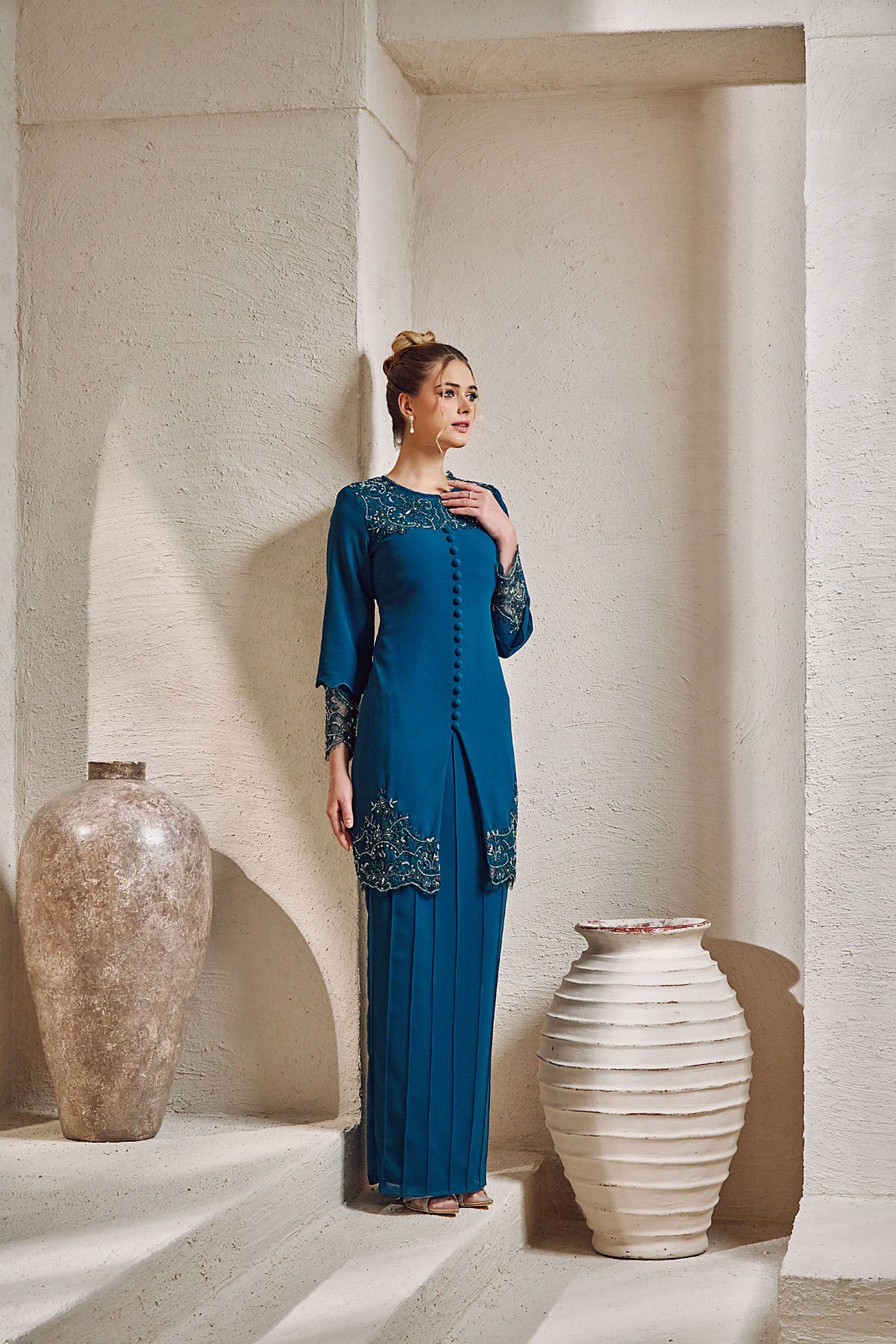 Woman in teal embroidered baju kurung with pleated skirt posing in arched stone setting.