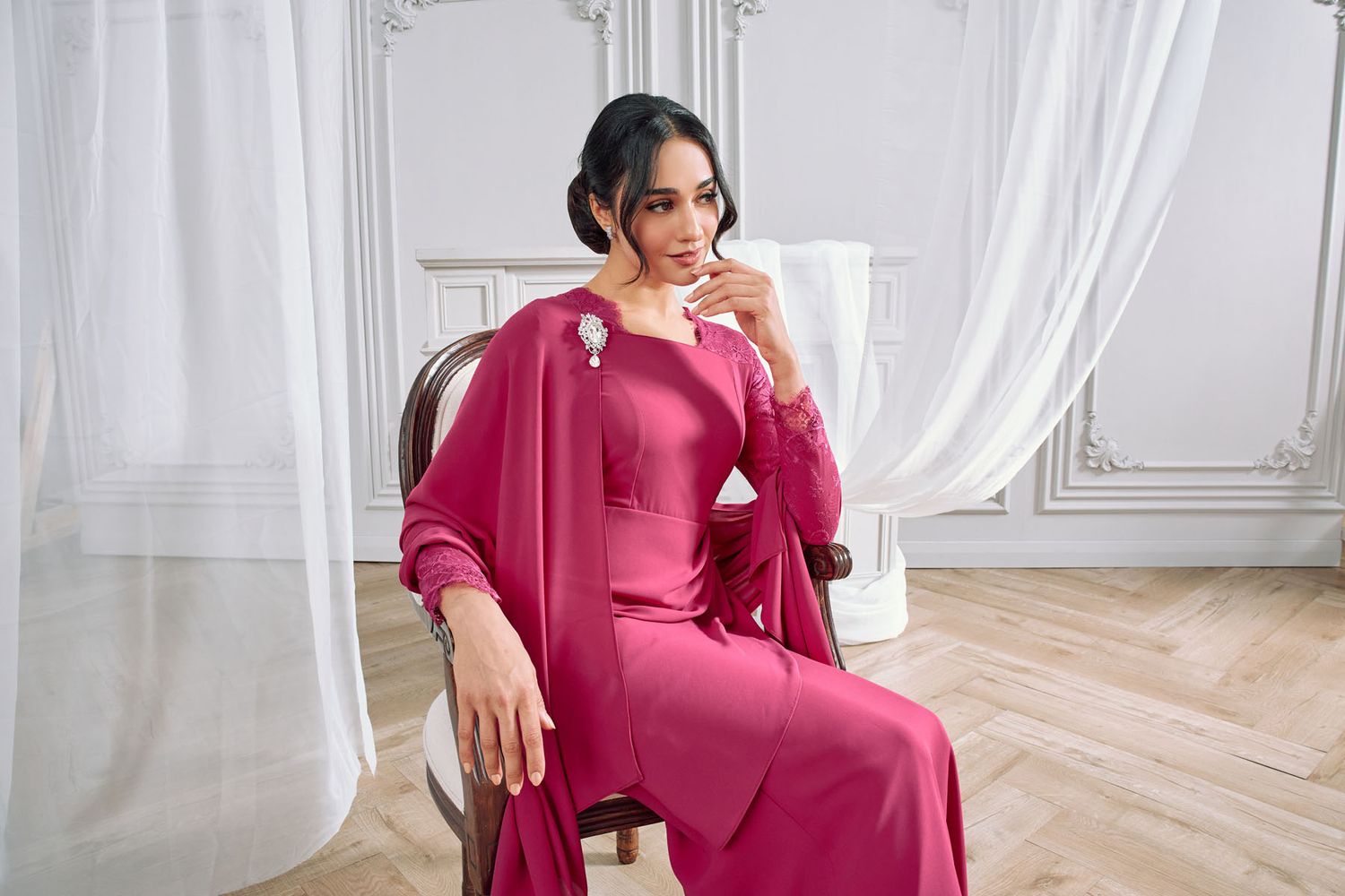 Woman in elegant fuchsia pink long dress seated on chair in a bright white classical interior.