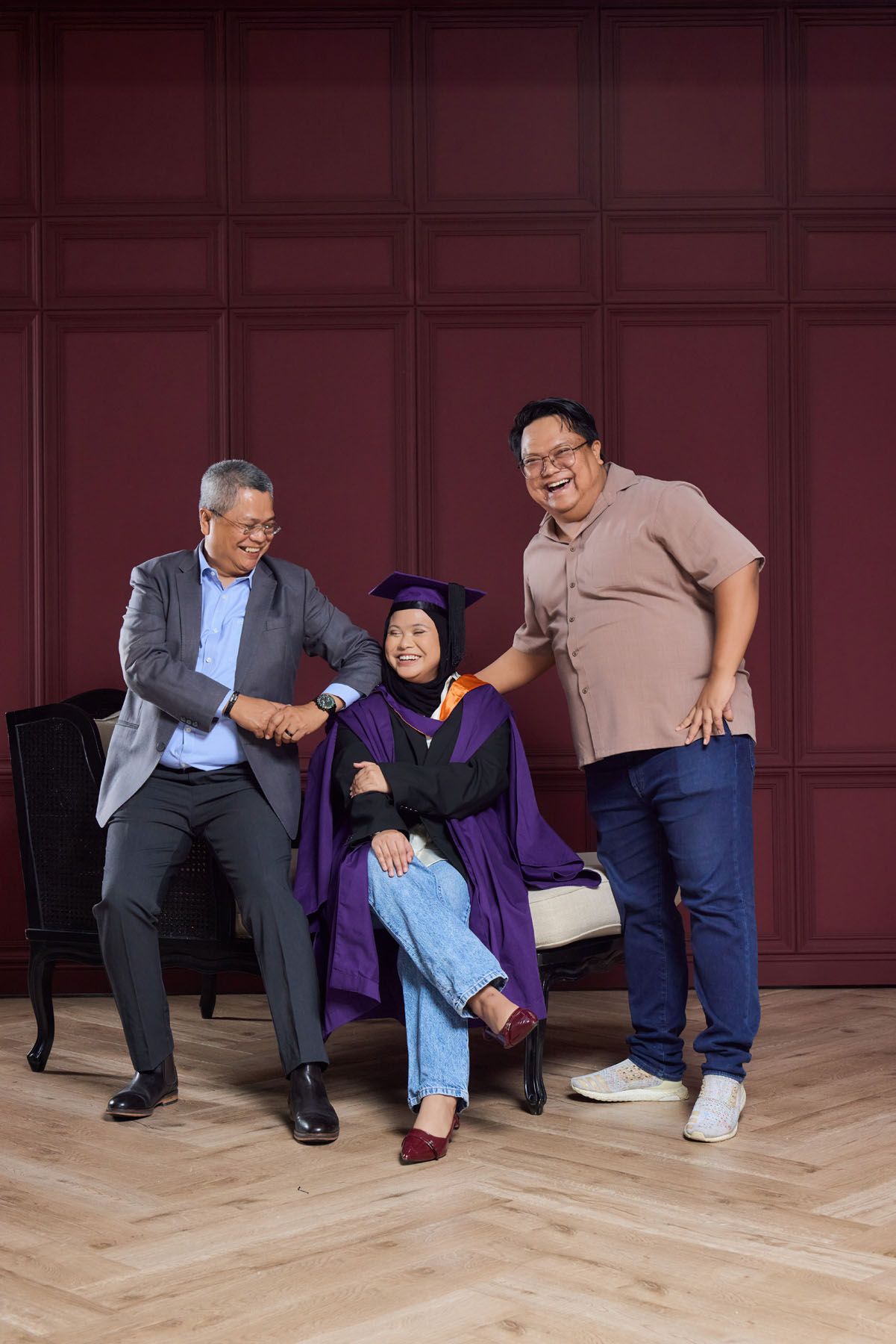 Graduate in purple gown seated between father and brother smiling in red-walled studio.