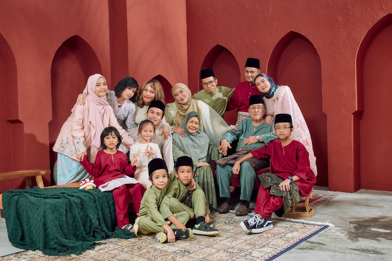 Large Malaysian family group portrait in colorful traditional Eid festive attire against red wall.