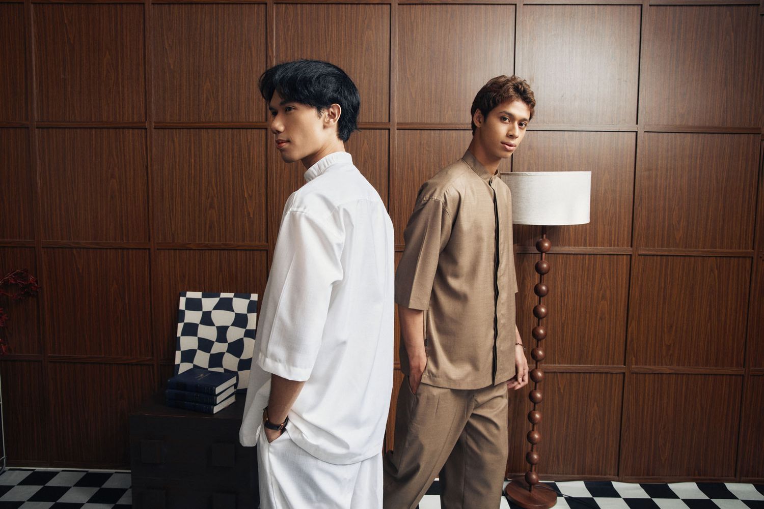 Two young men in white and tan modern baju melayu sets stand back-to-back in stylish room.