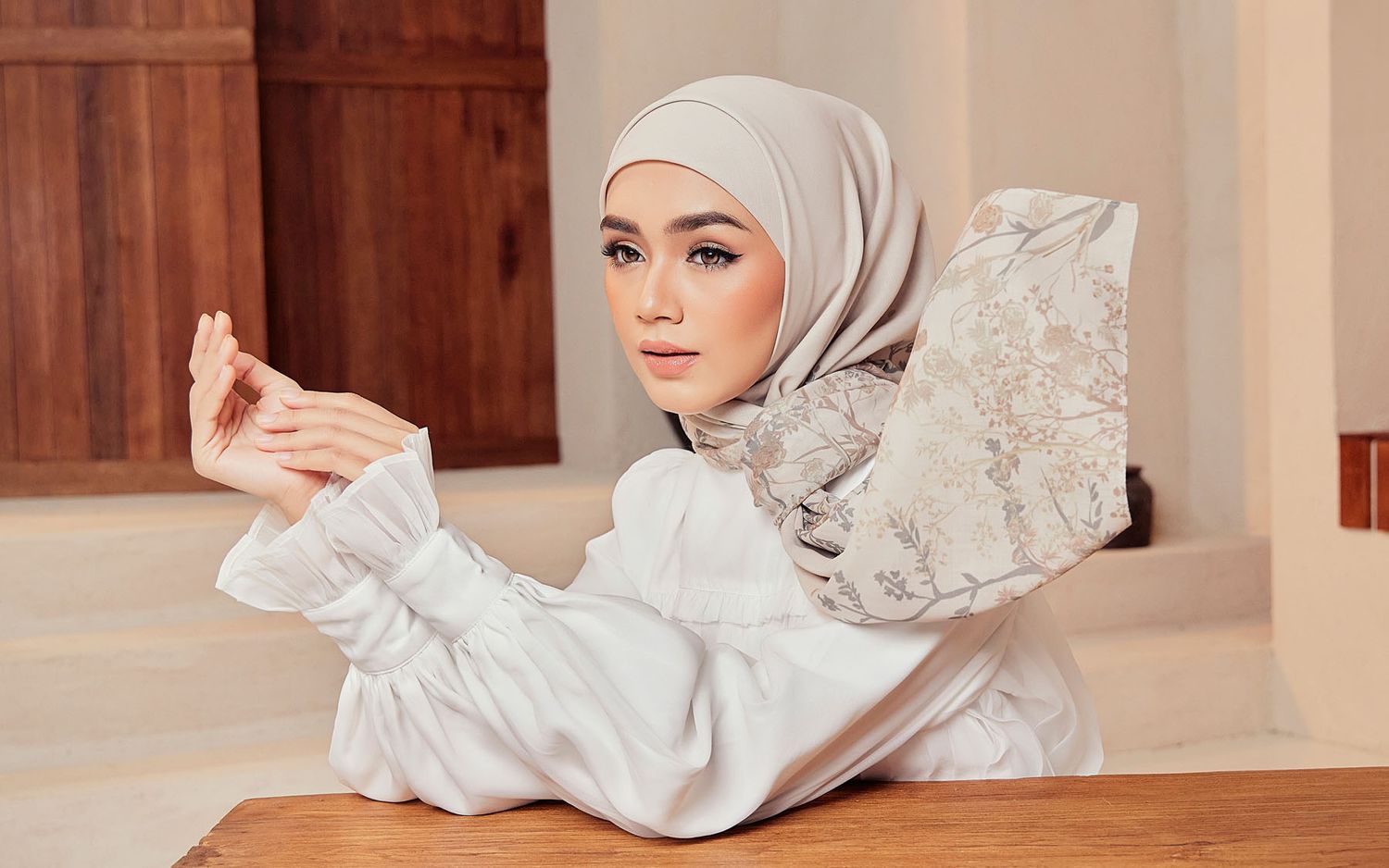 Stylish woman in a grey hijab and white lace blouse posing with hands raised in a serene setting.