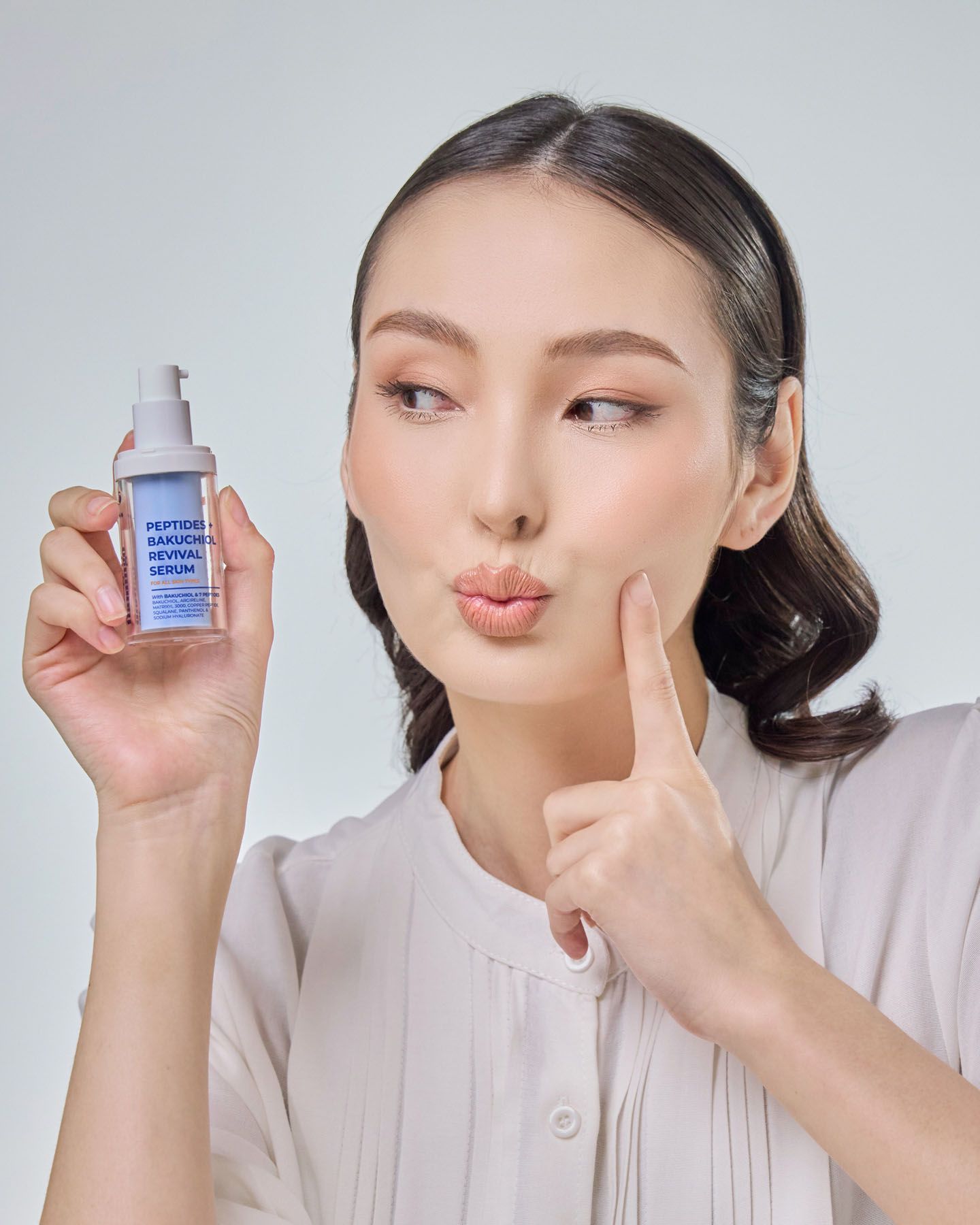 Woman pointing to cheek holding Peptides Niacinamide Revival Serum bottle, making kissing face.