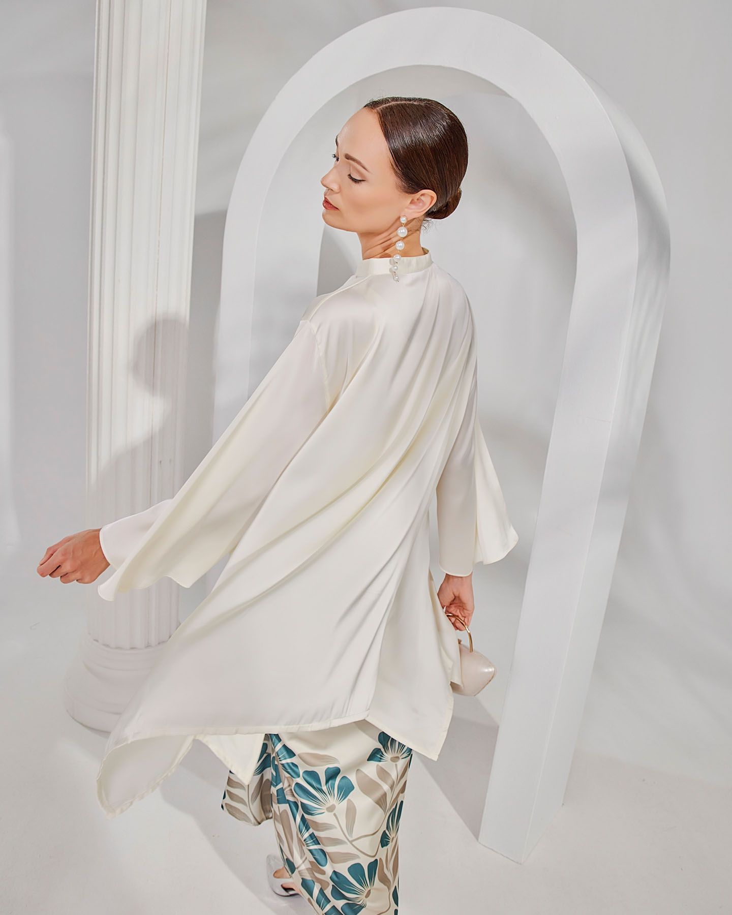A woman in a flowing white satin kaftan top and floral pants poses gracefully near a white arch.
