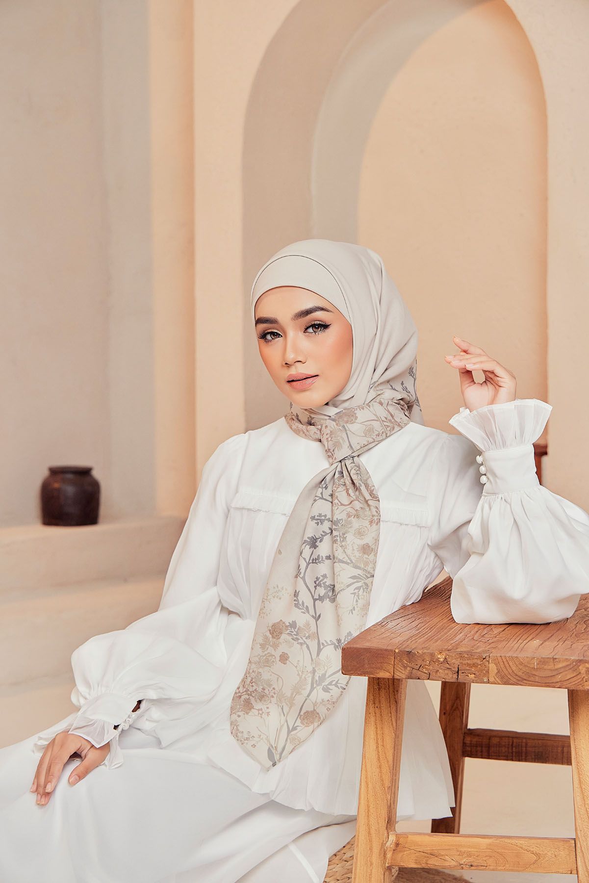 A woman in a white dress and beige hijab wears a floral printed scarf while seated on a wooden bench indoors.