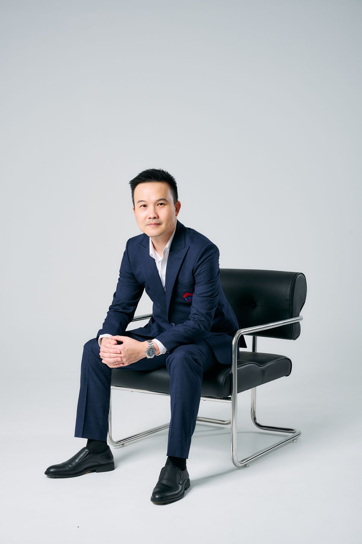 Professional Asian businessman in navy suit sitting confidently on black leather chair against gray background.