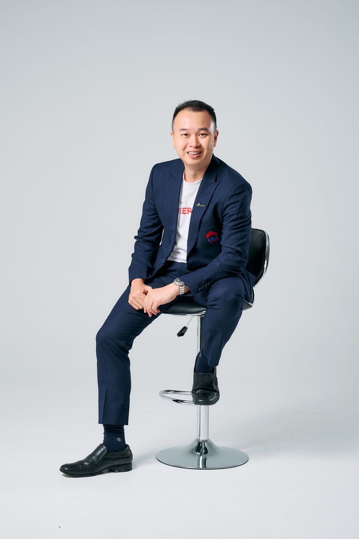 A smiling Asian man in a navy blue suit sits confidently on a bar stool against a light gray background.