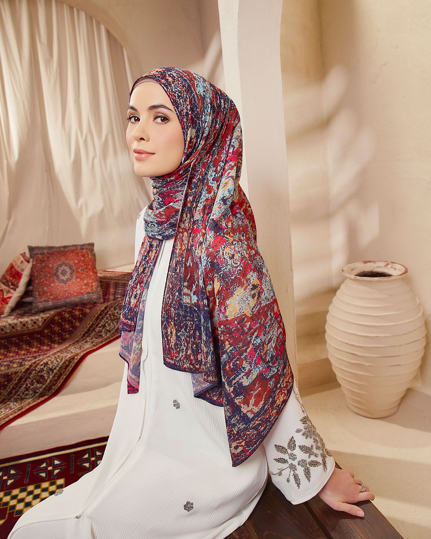 Woman in white outfit wearing colorful patterned red hijab seated in elegant Middle Eastern inspired setting.