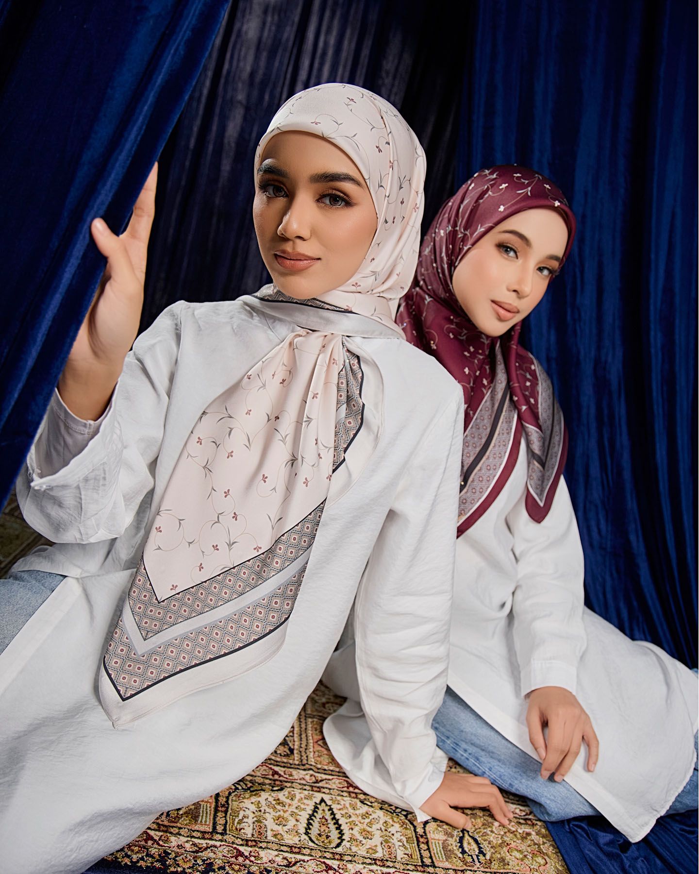 Two women in white outfits wearing patterned hijabs posing together against a blue curtain.