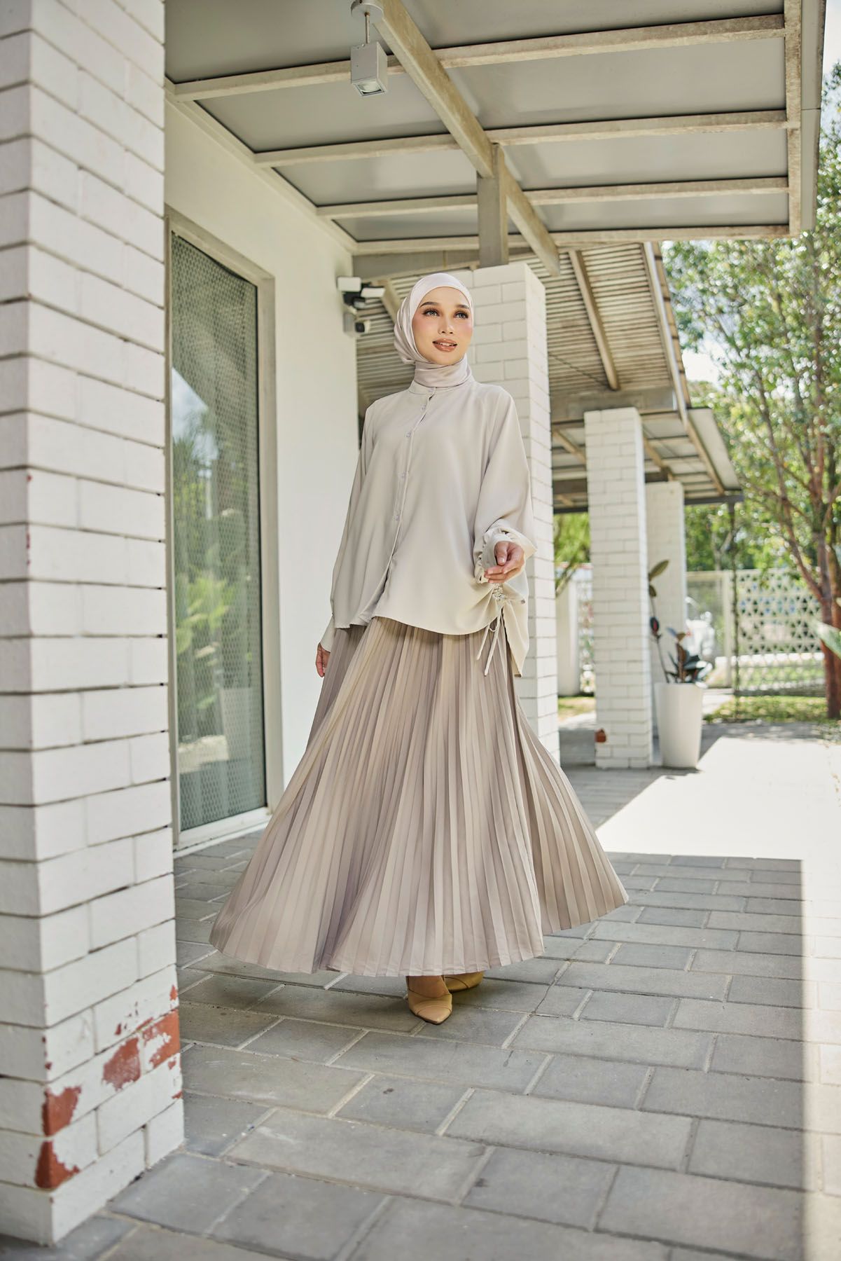 A hijab-wearing woman in a beige pleated skirt and matching top walks along a sunlit outdoor corridor.