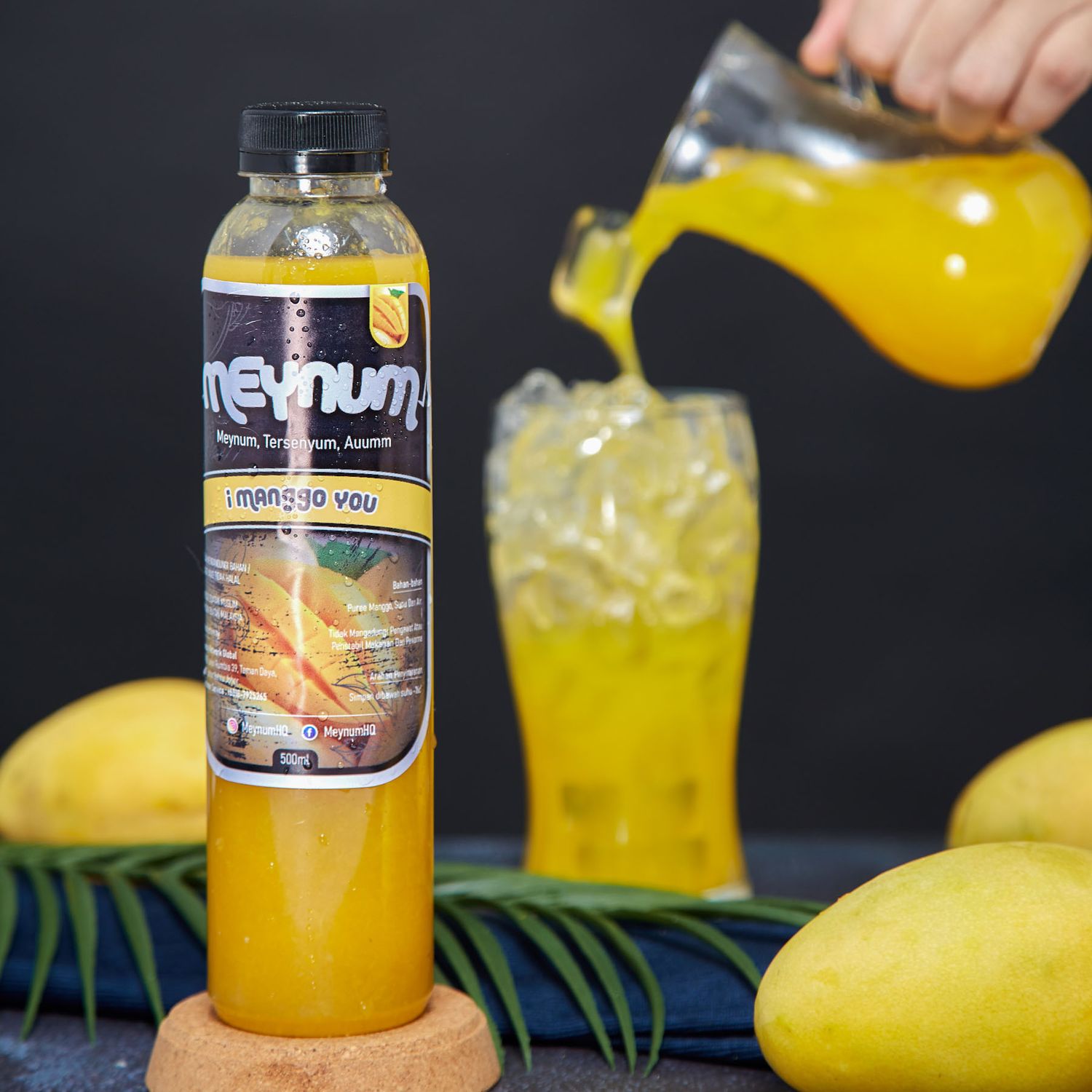 A bottle of Meynum mango juice being poured into a glass with fresh mangoes and tropical leaves nearby.