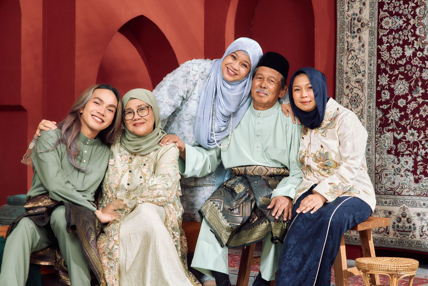 Family of five in coordinating sage green and cream traditional Malay outfits posing against red arched wall.