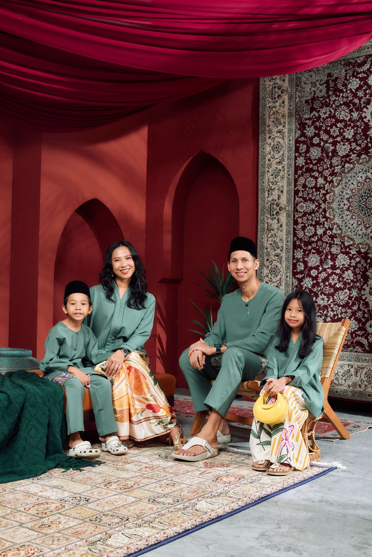 Family of four wearing sage green traditional Malaysian outfits seated in a vibrant red Moroccan-style setting.