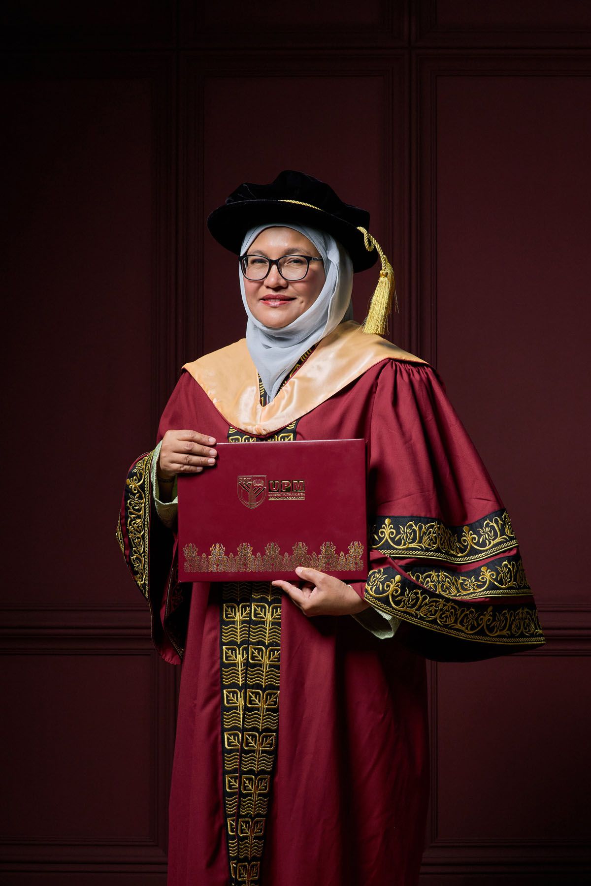Woman in maroon doctoral graduation gown and hijab holding diploma certificate at university convocation ceremony.