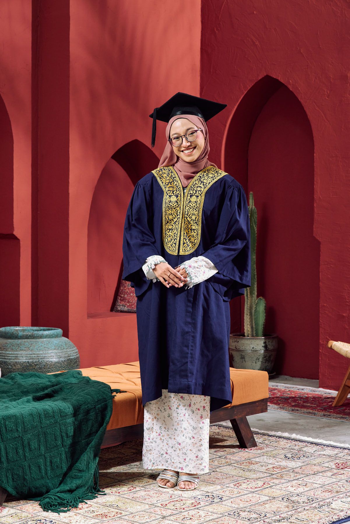 Smiling Muslim graduate in navy blue gown and hijab stands before a vibrant red Moroccan archway.
