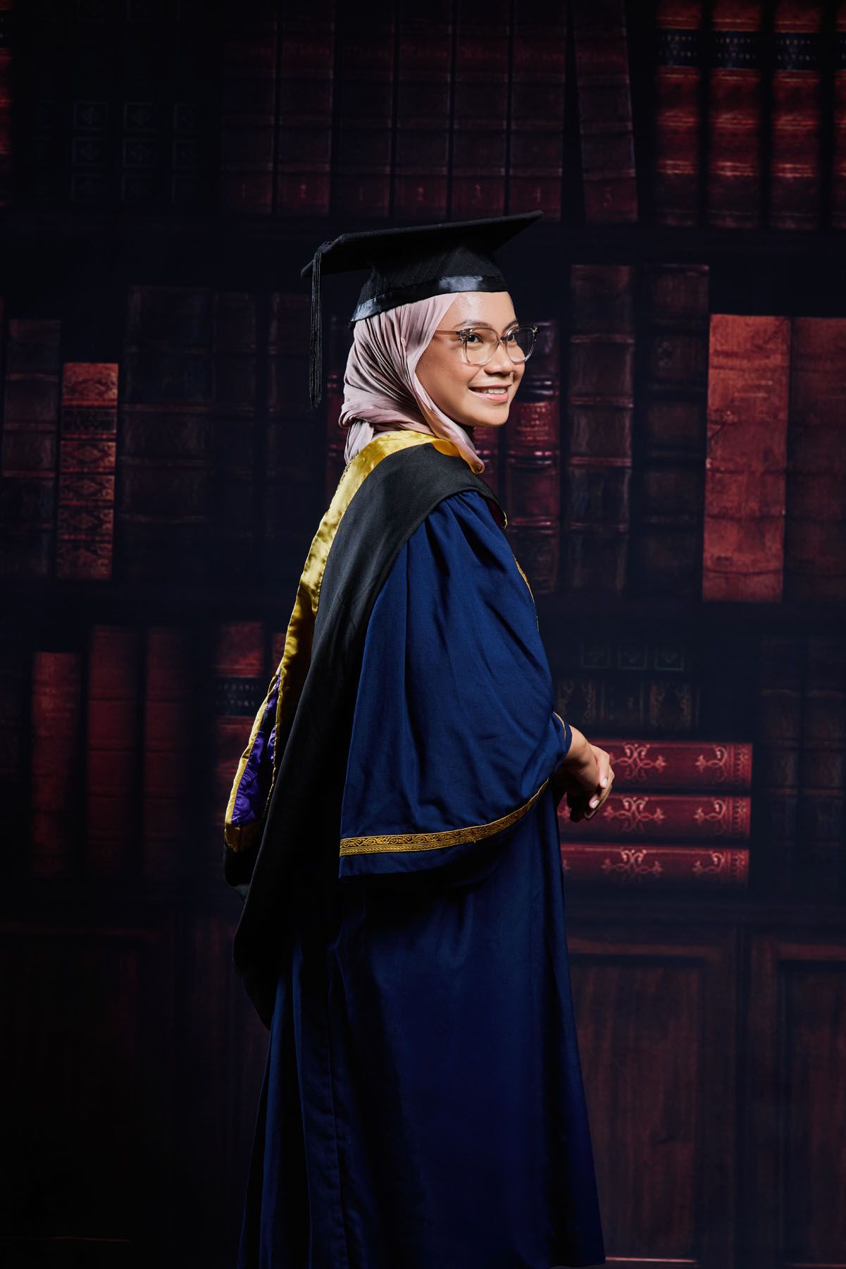 Female Muslim graduate in navy robe posing before a bookshelf backdrop.
