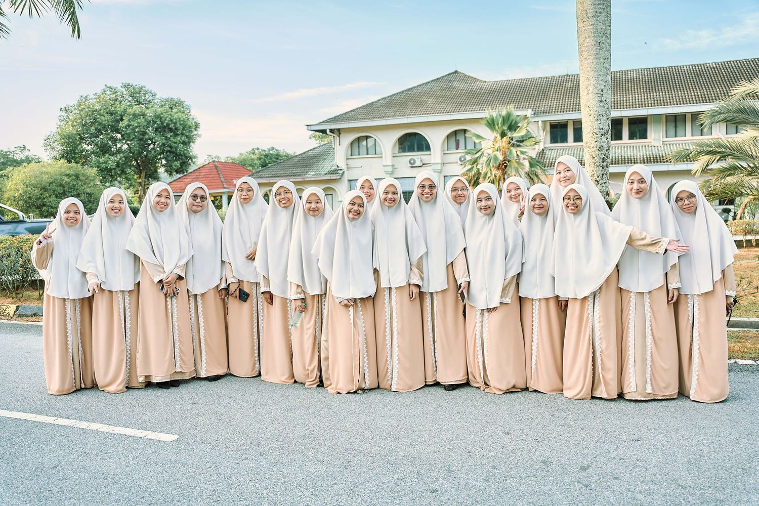 Large group of women in matching white and beige hijab and jubah posing outdoors.